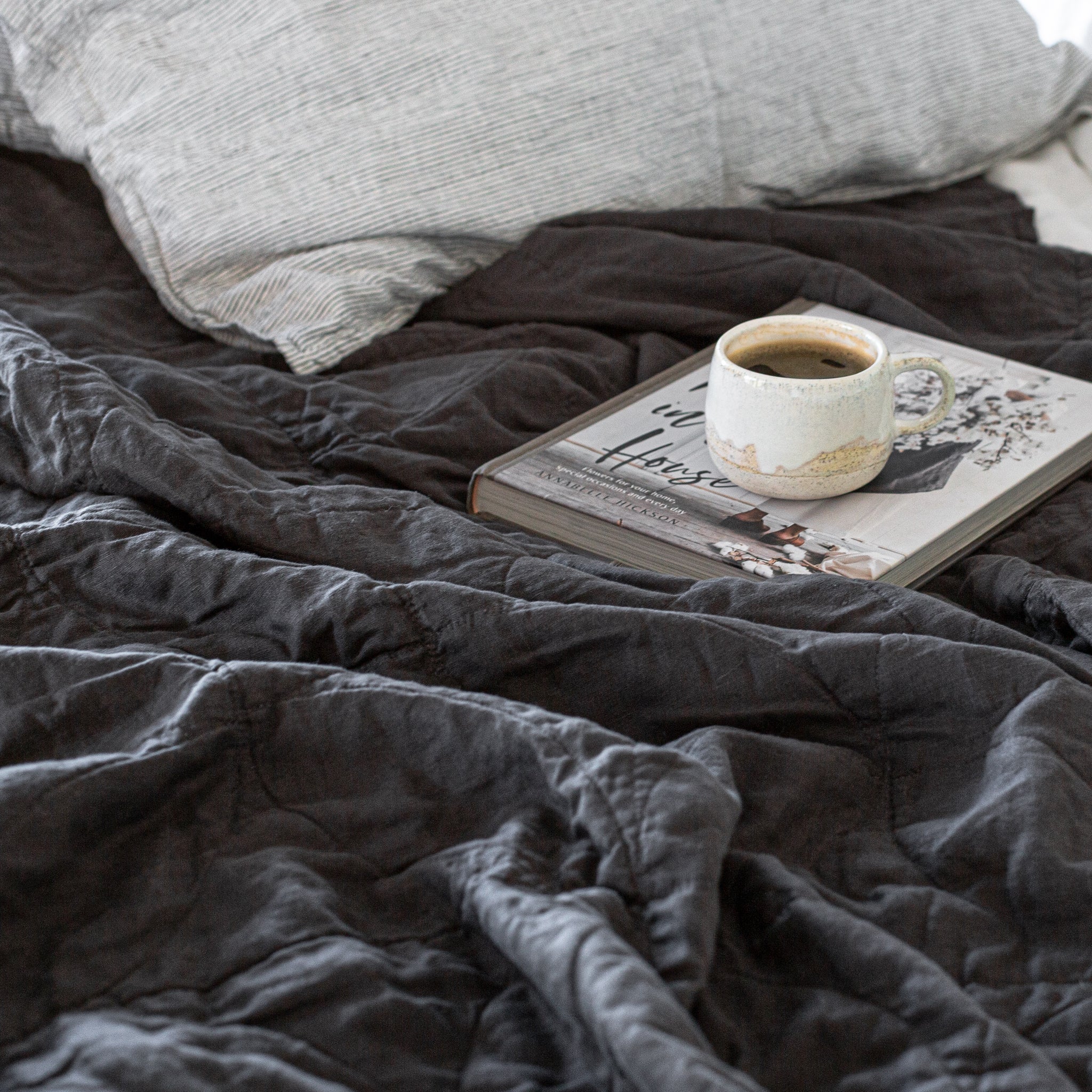 A dark charcoal linen blanket at the foot of a bed in a room with a white wall and a lamp.