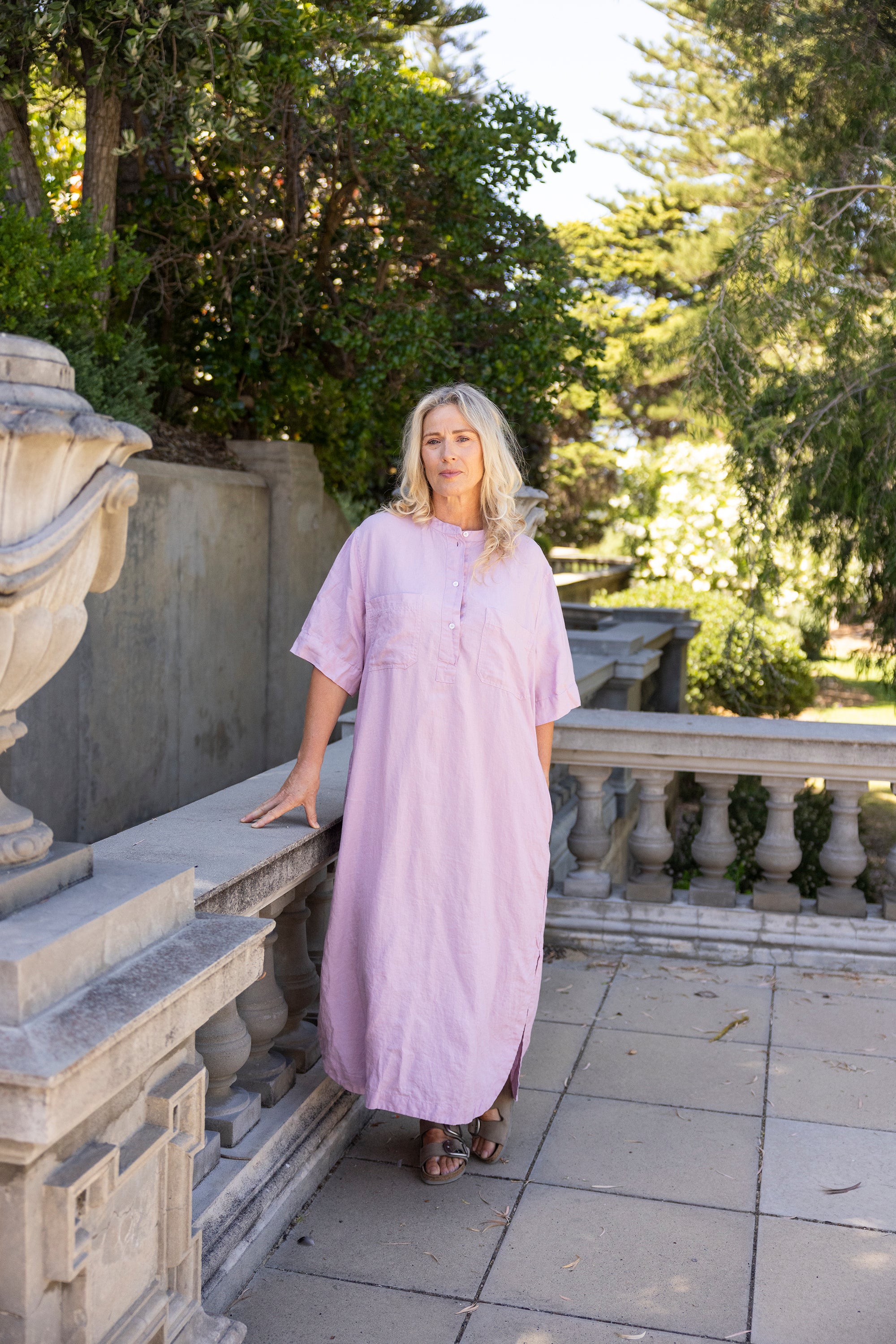 Woman in a pink dress standing in a garden setting with trees and stone structures.