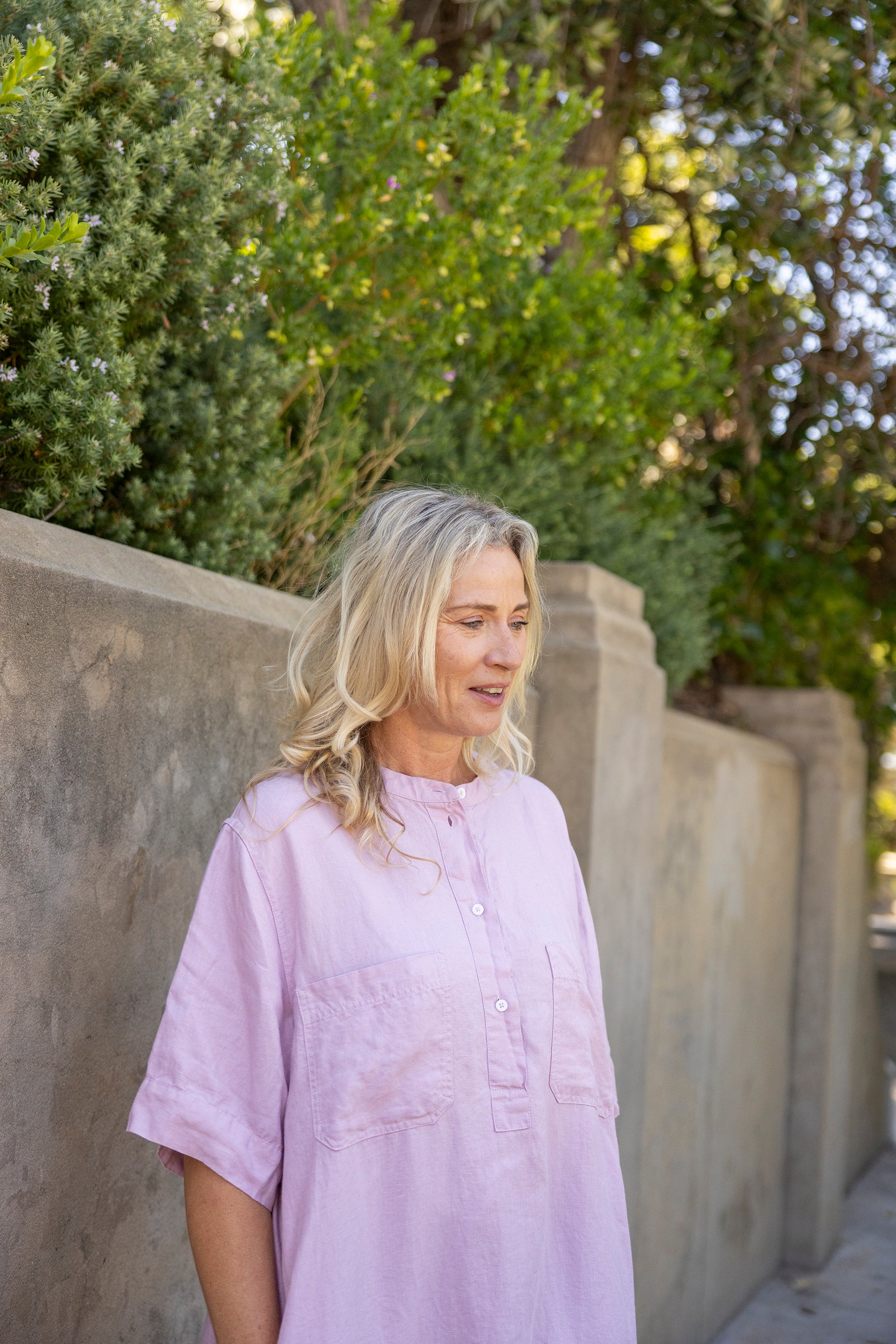 Woman in a light purple shirt standing against a stone wall with greenery in the background