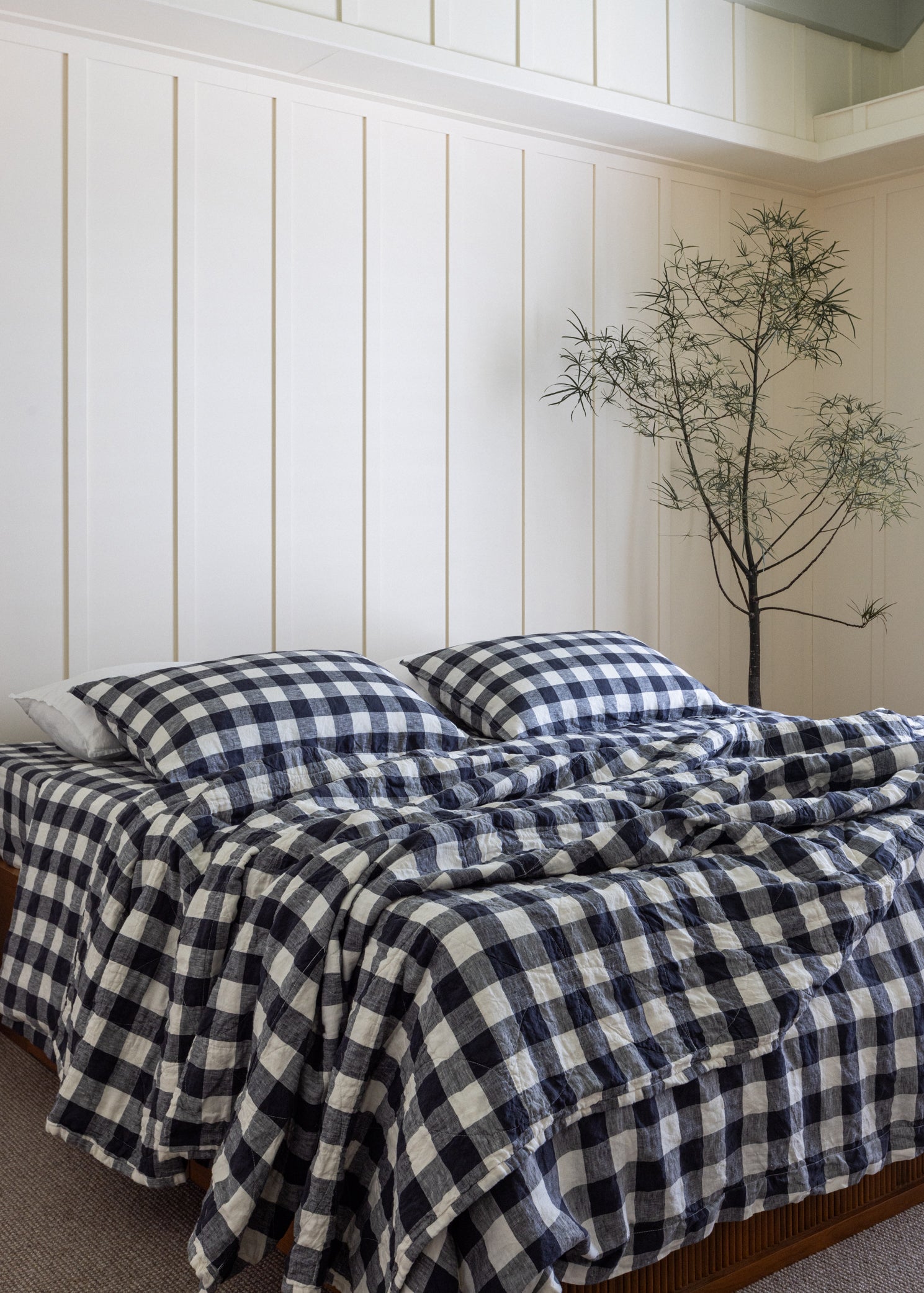 A bedroom setting with a bed covered by a blue and white checkered linen blanket, circle stitched, placed against a white wall with vertical paneling.