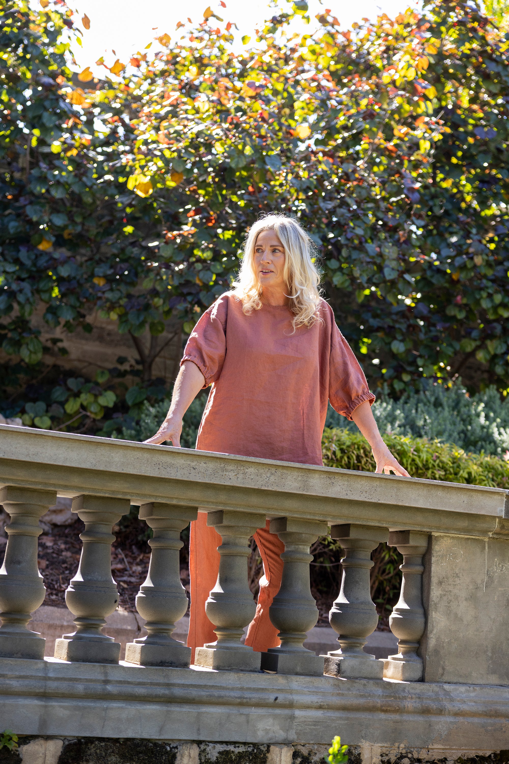 Woman standing on a stone railing with greenery in the background wearing rust-coloured oversized boxy top