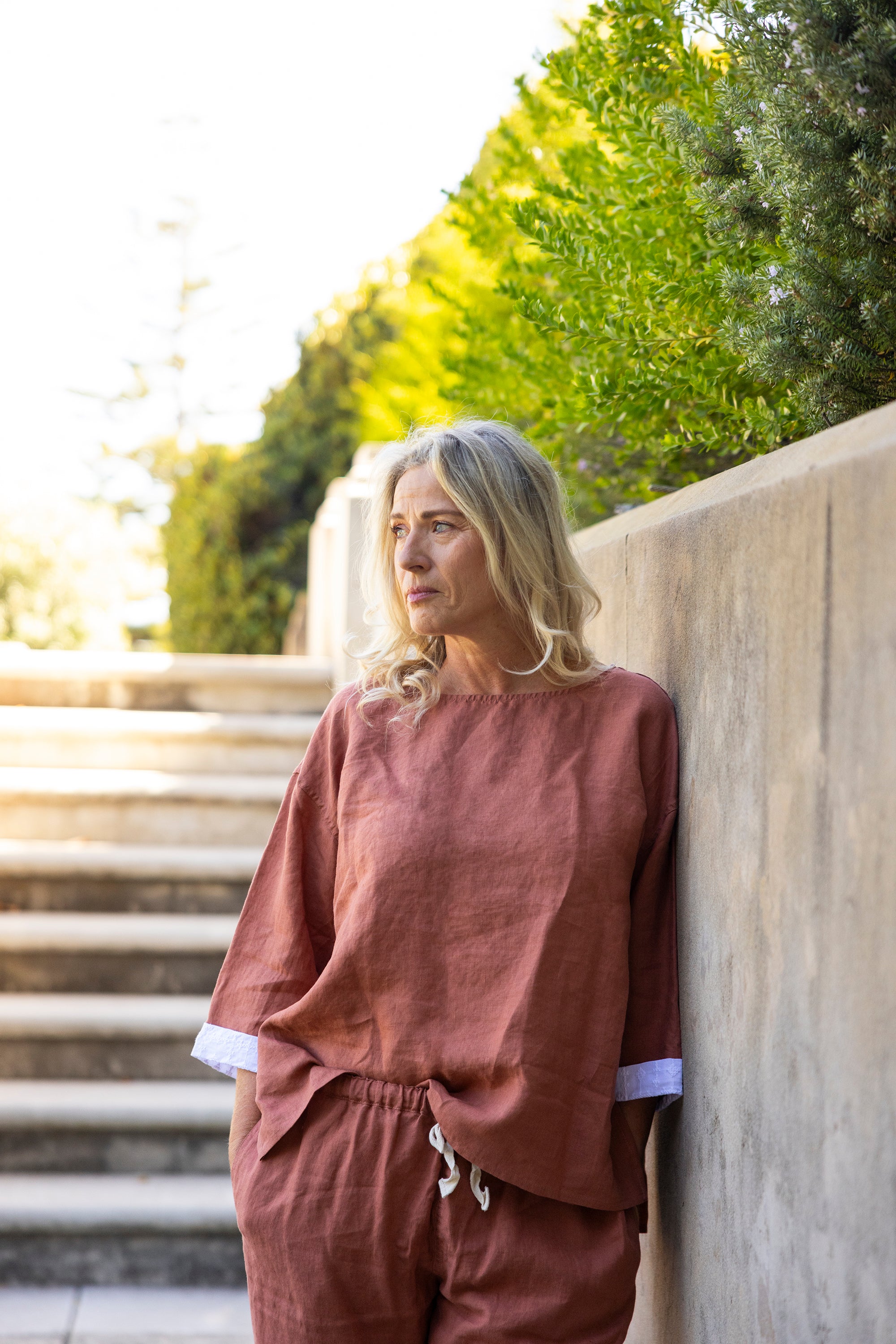Woman in an oversized relaxed-fit boxy top with contrasting sleeve hem in a rust colour leaning against a stone wall with greenery in the background