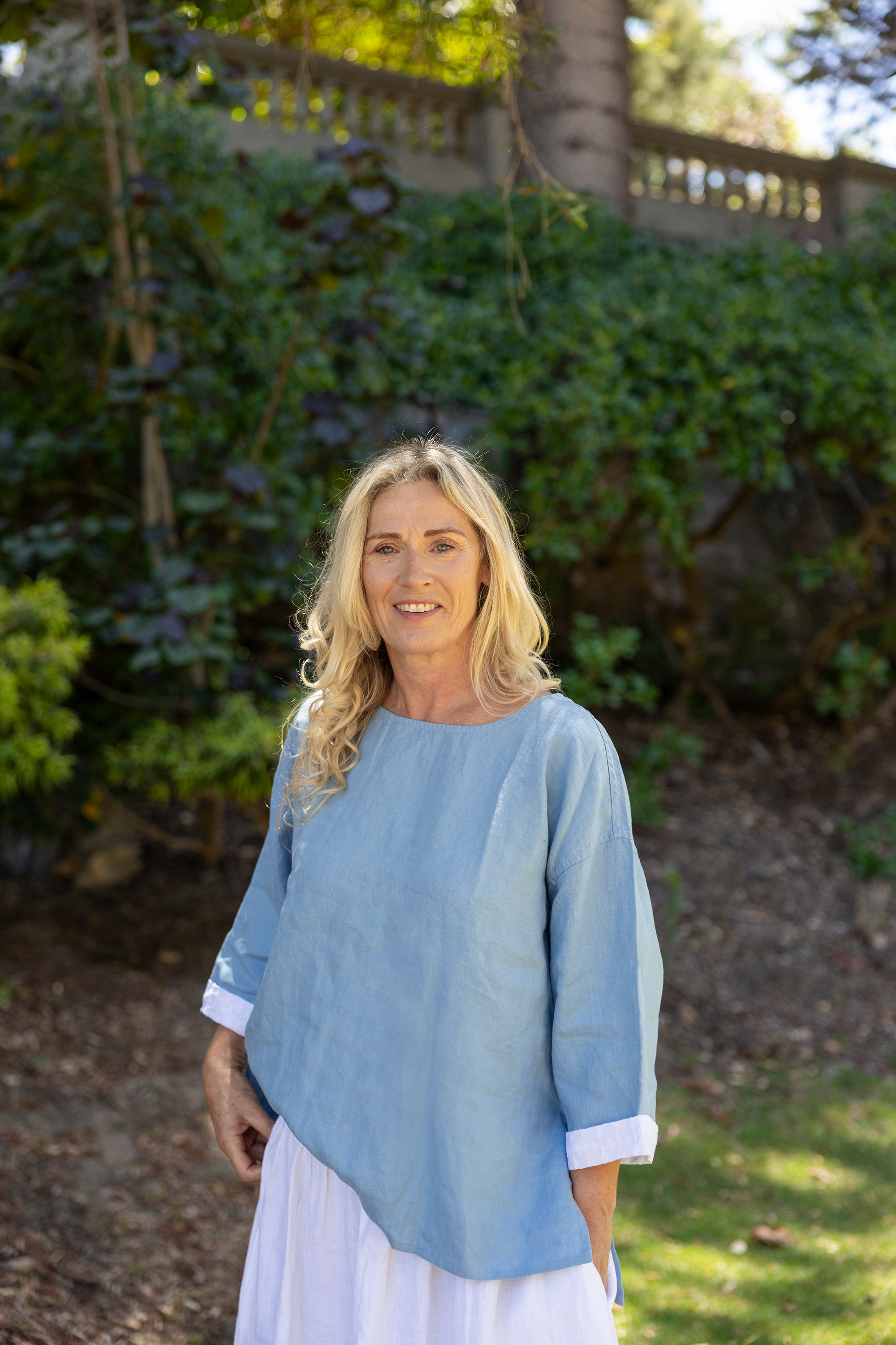 Woman wearing a light blue top and white skirt standing in a garden.
