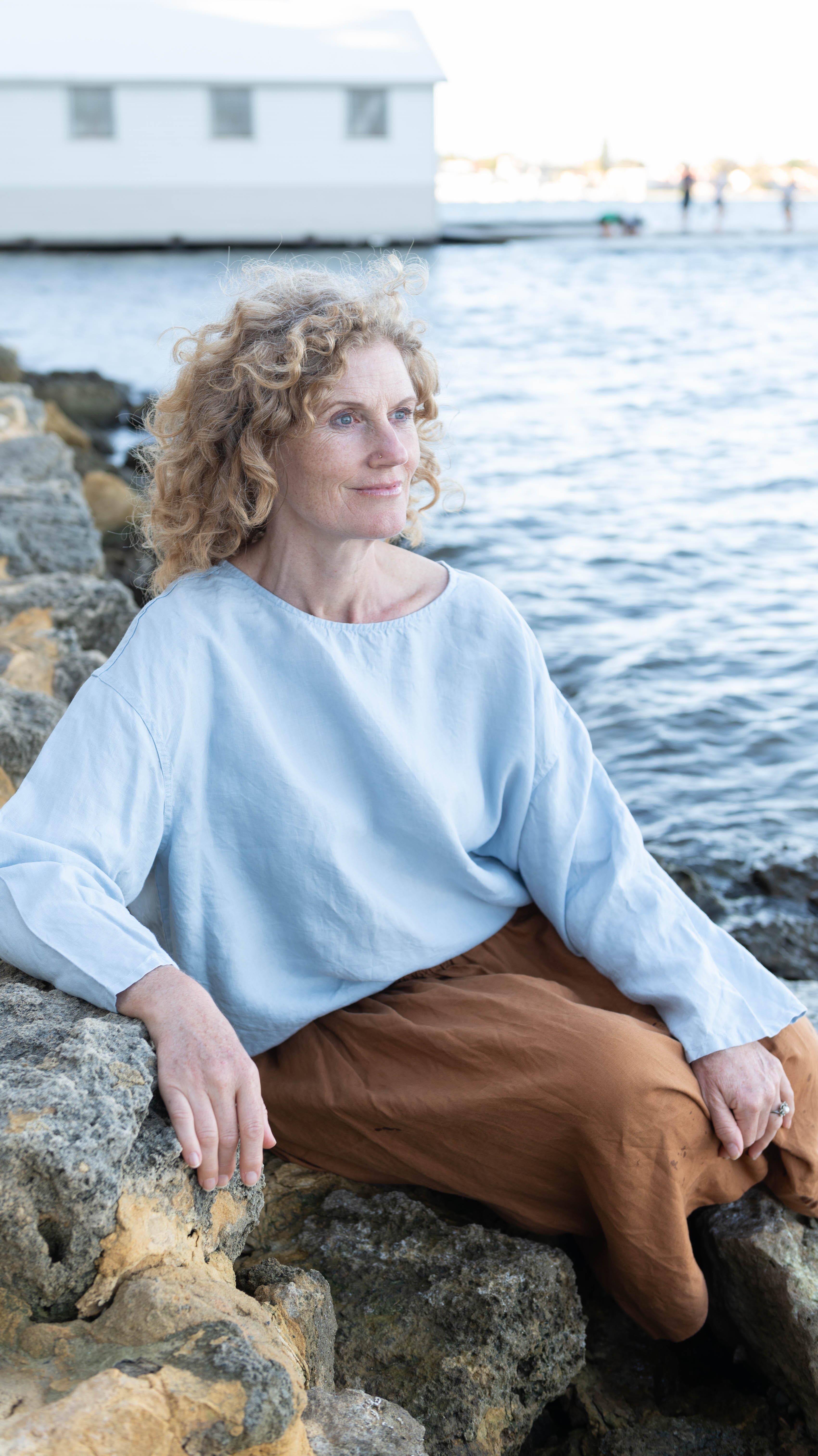 A woman seated outdoors wearing a relaxed fit, long-sleeved Boxy Top in a pale blue color, paired with a terracotta skirt.