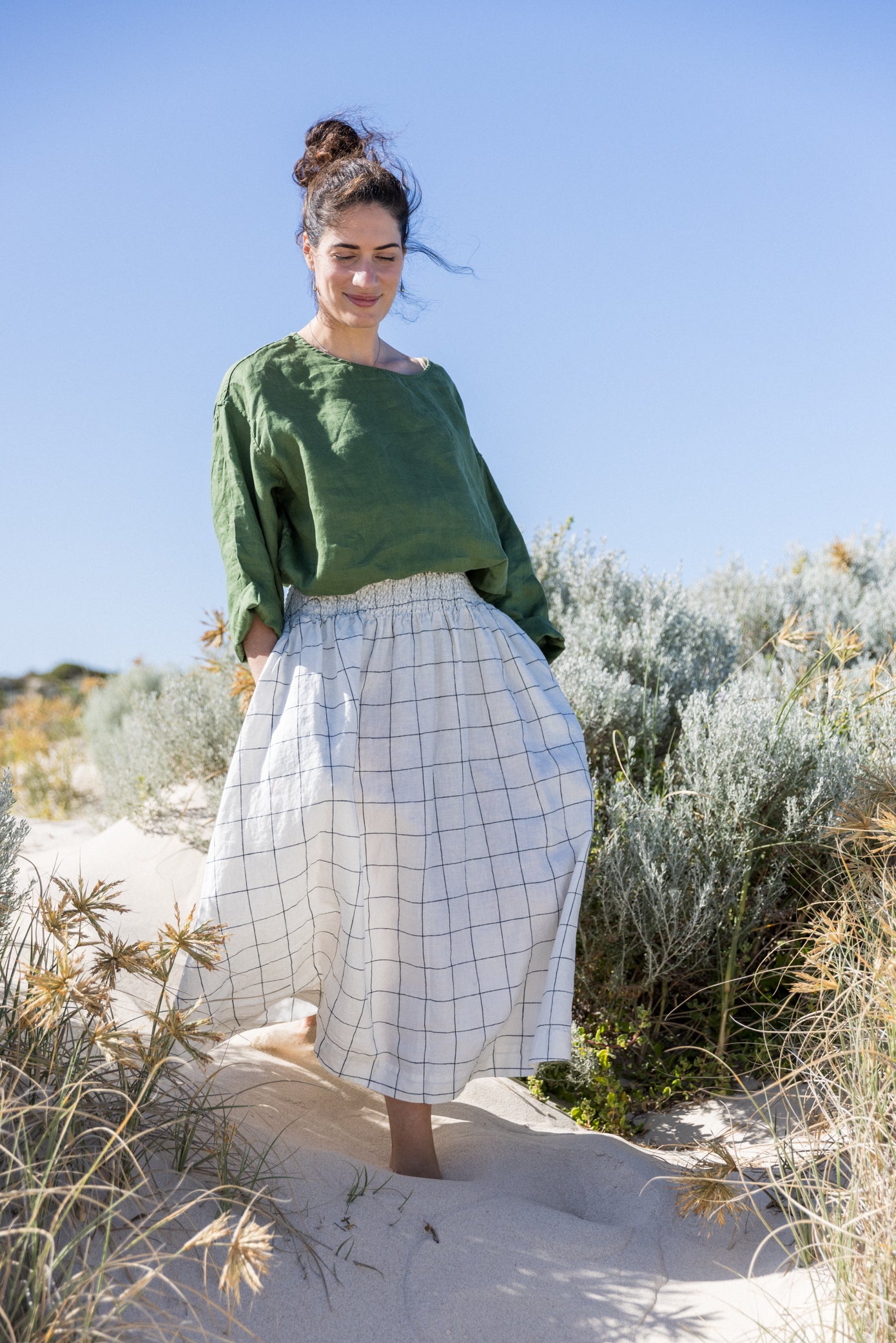 A woman standing outdoors on the beach wearing a relaxed fit, long-sleeved Boxy Top in a dark green color, paired with a grid-patterned skirt.