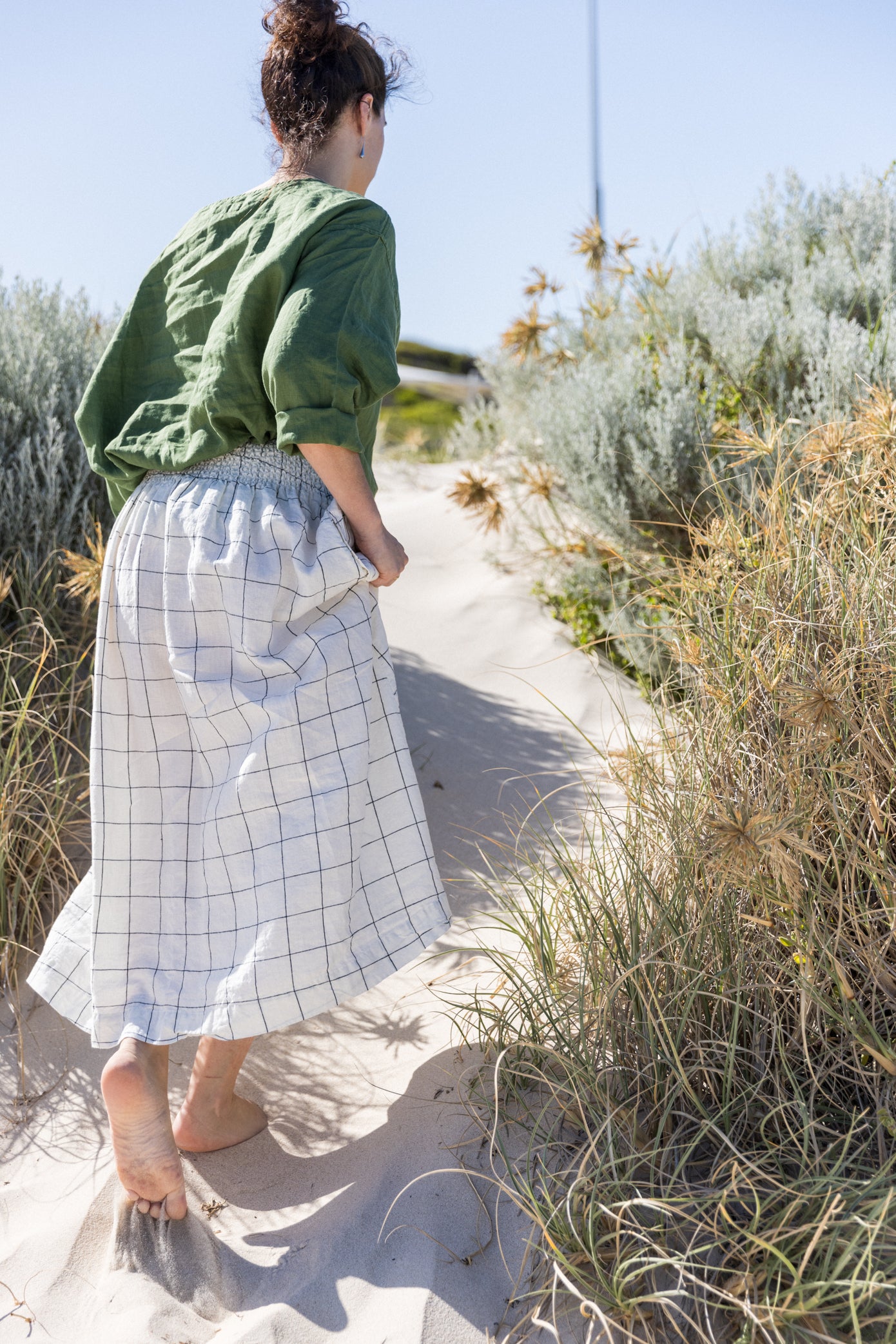 Woman standing outdoors on a beach wearing a relaxed-fit boxy top in a dark green colour paired with a grid-patterned skirt