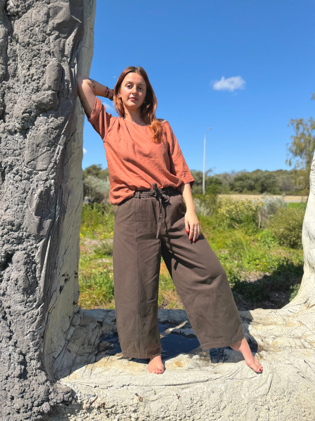 A woman stands outdoors between two large stone pillars, wearing a rust-orange short-sleeve top and wide-leg brown pants. The background features a bright blue sky, green grass, and distant trees.