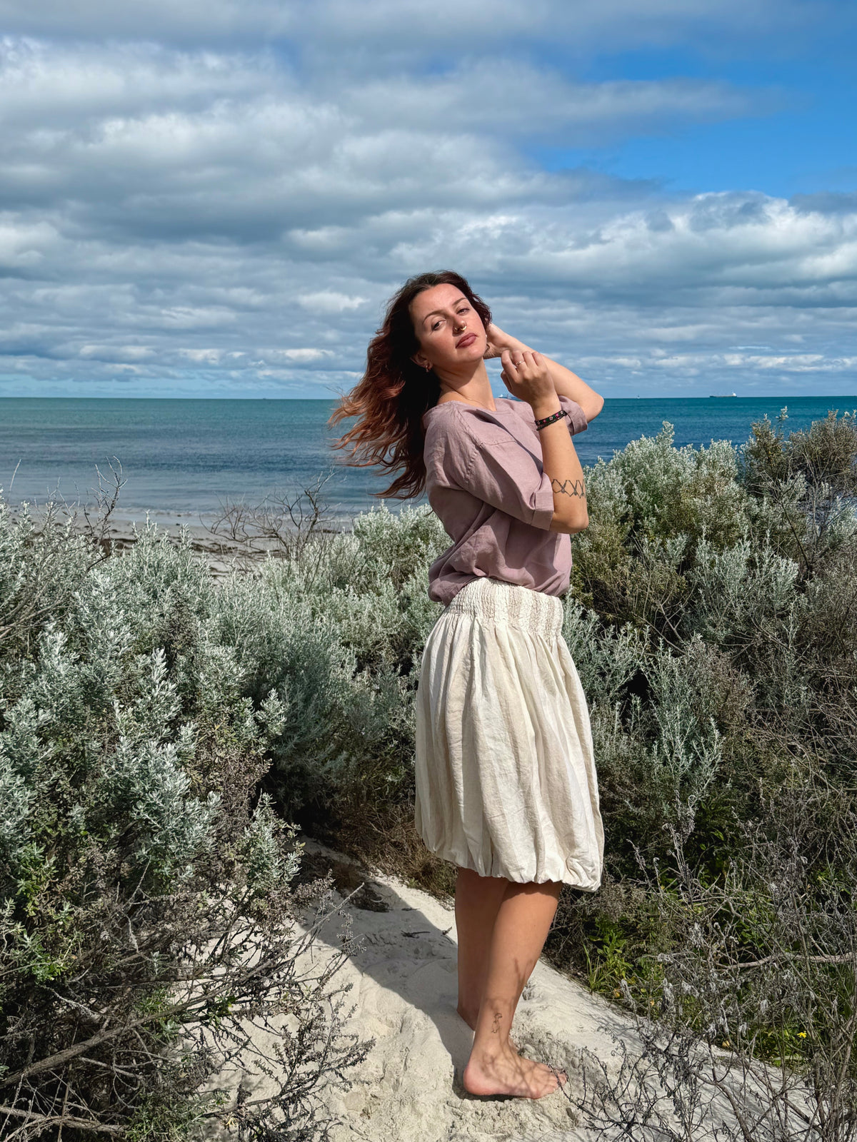 A woman stands barefoot on a sandy path outdoors by the ocean, surrounded by green shrubs, wearing a mauve-brown short-sleeve top and a cream pleated skirt. The background features a blue ocean and a cloudy sky.
