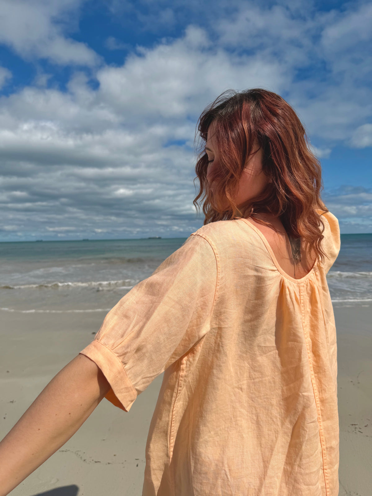 A woman with reddish-brown hair stands on a sandy beach facing the ocean under a blue sky with white clouds, wearing a loose, short-sleeve peach-colored linen top.