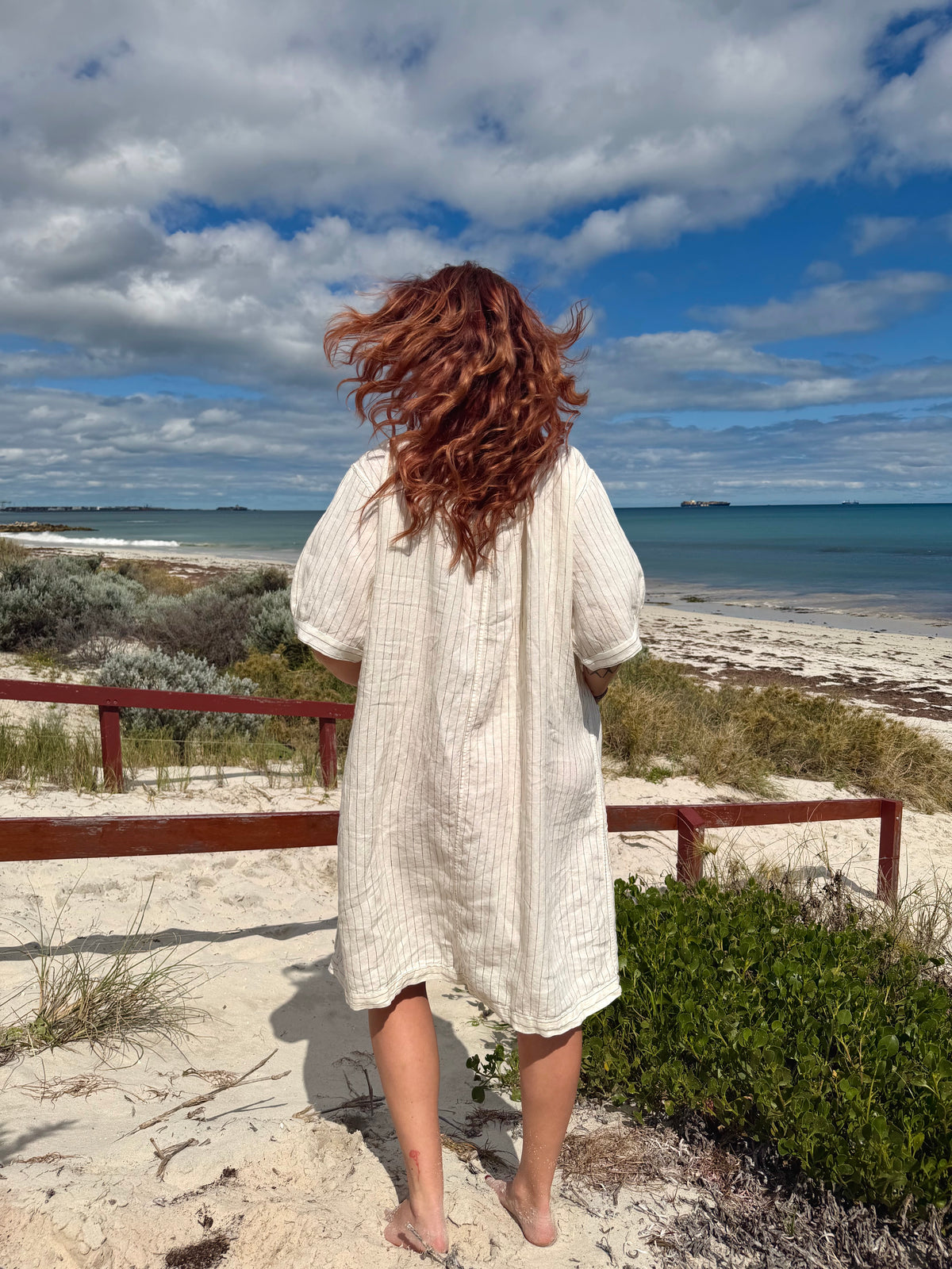 A woman with long, wavy auburn hair stands on a sandy beach path, viewed from behind, wearing a cream-colored textured linen-blend tunic or cover-up dress. The background features a scenic view of the ocean, blue sky with clouds, and green beach vegetation.