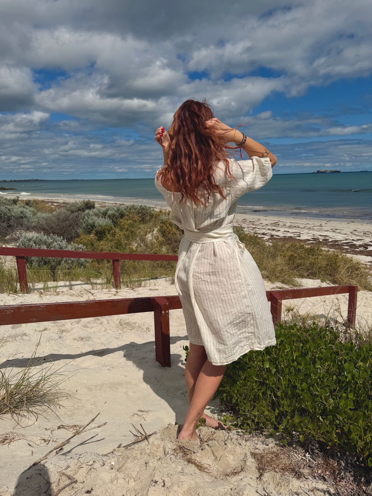 A woman stands on a sandy beach dune with a wooden bench, looking out towards the ocean under a cloudy blue sky. She is wearing a cream-colored, textured linen wrap dress.