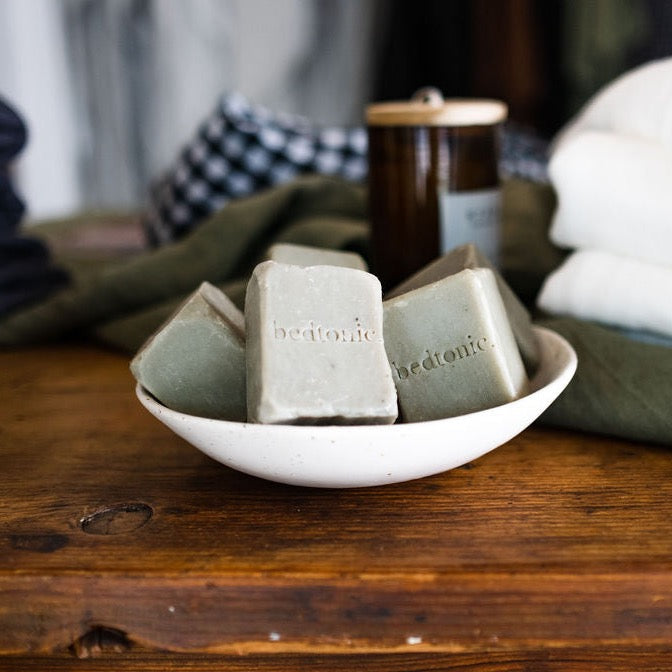 Three bars of soap with the text 'bedtonic' inscribed on them, placed in a white bowl on a wooden surface, with a blurred background featuring a checkered pattern and a wooden object.