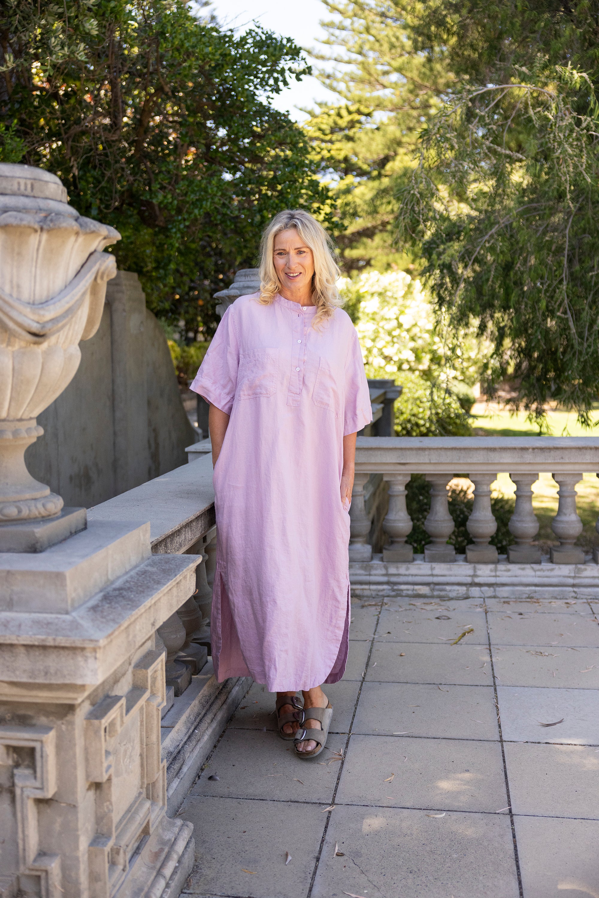 Woman in a long light purple dress standing on a stone pathway with greenery in the background