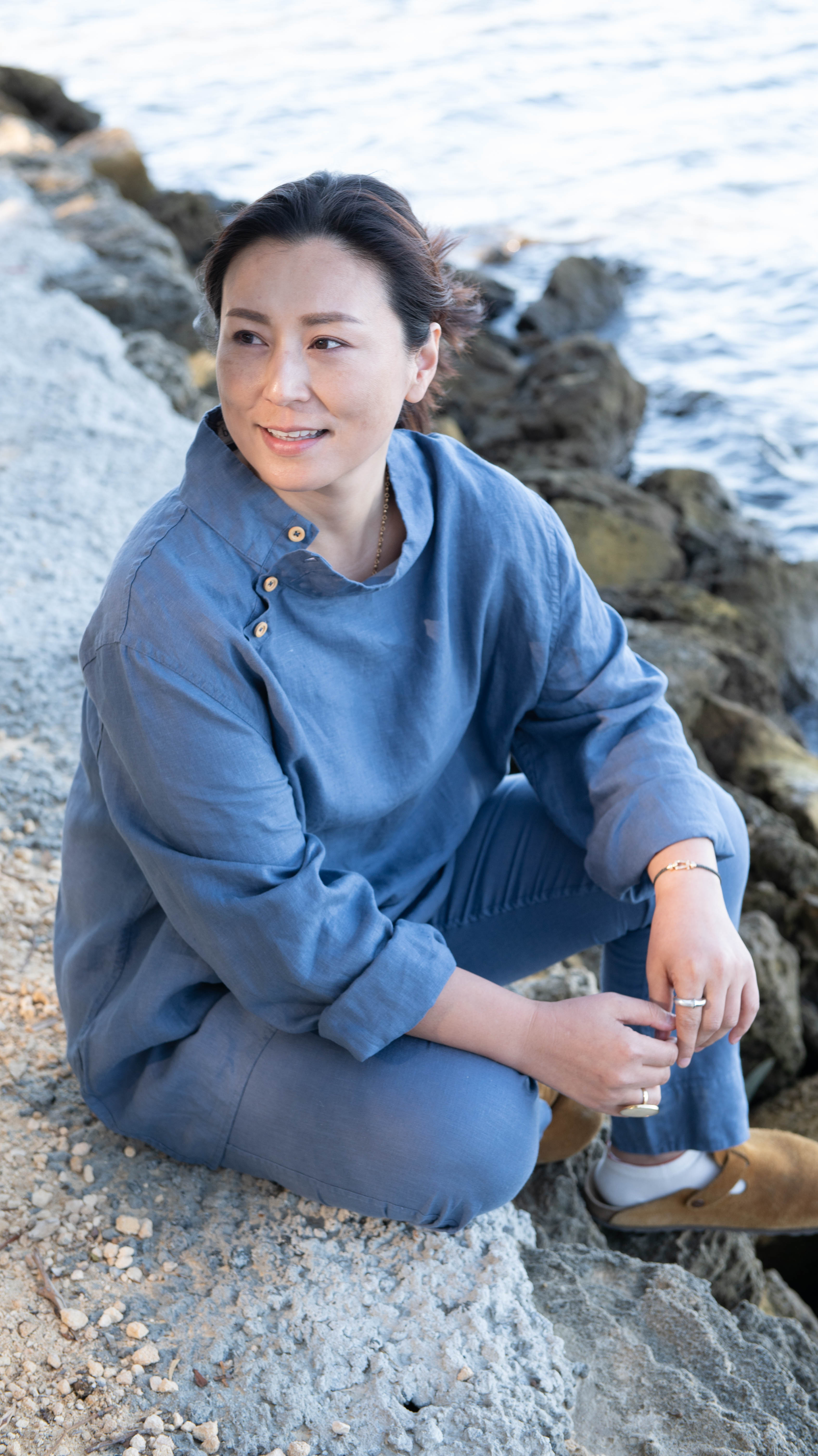 Woman in a denim blue tunic and matching trousers sat cross legged in front of a lake 