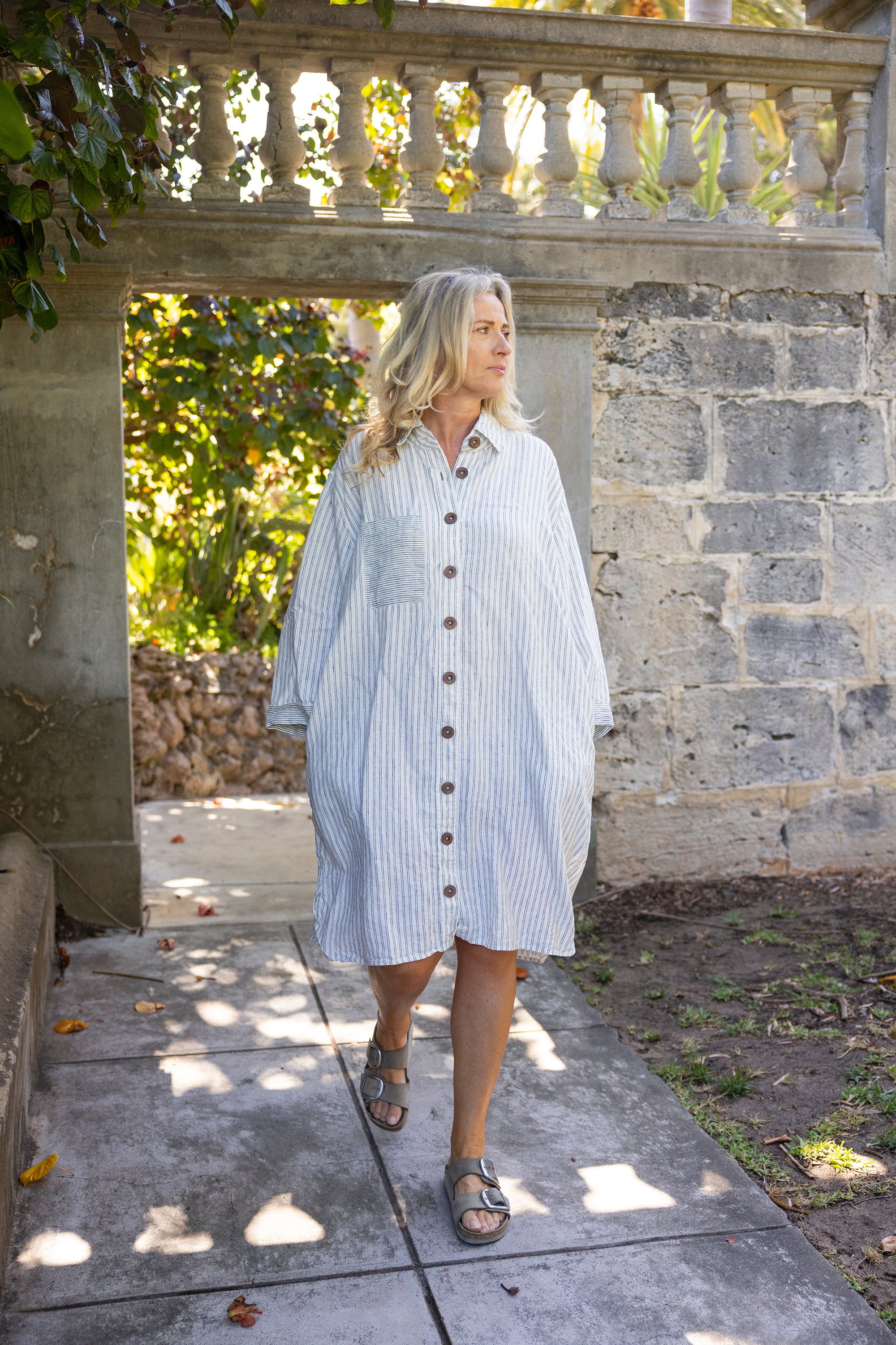 Woman wearing a blue and white striped dress walking outdoors near a stone wall.