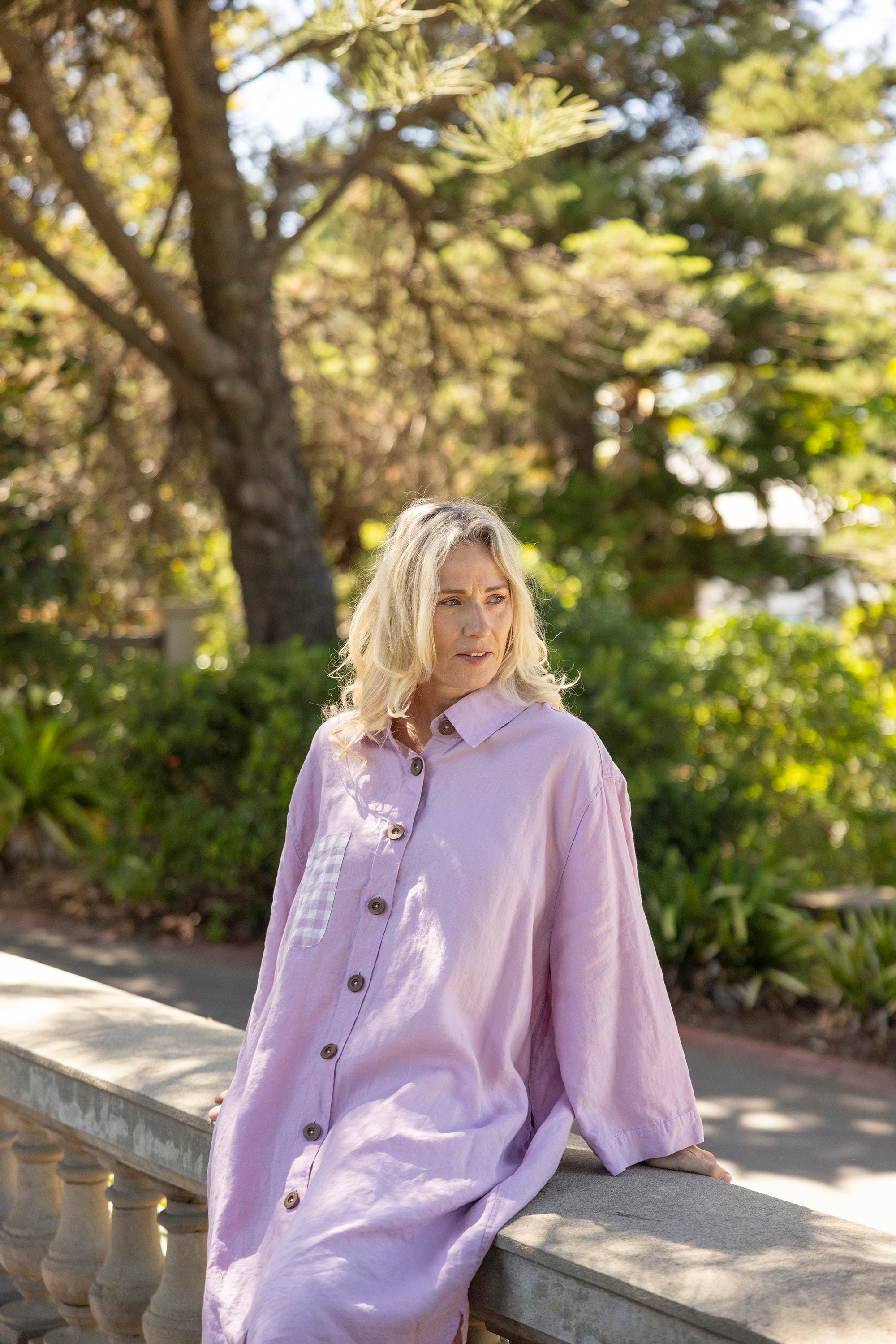 Woman in a light purple shirt sitting on a stone bench with greenery in the background