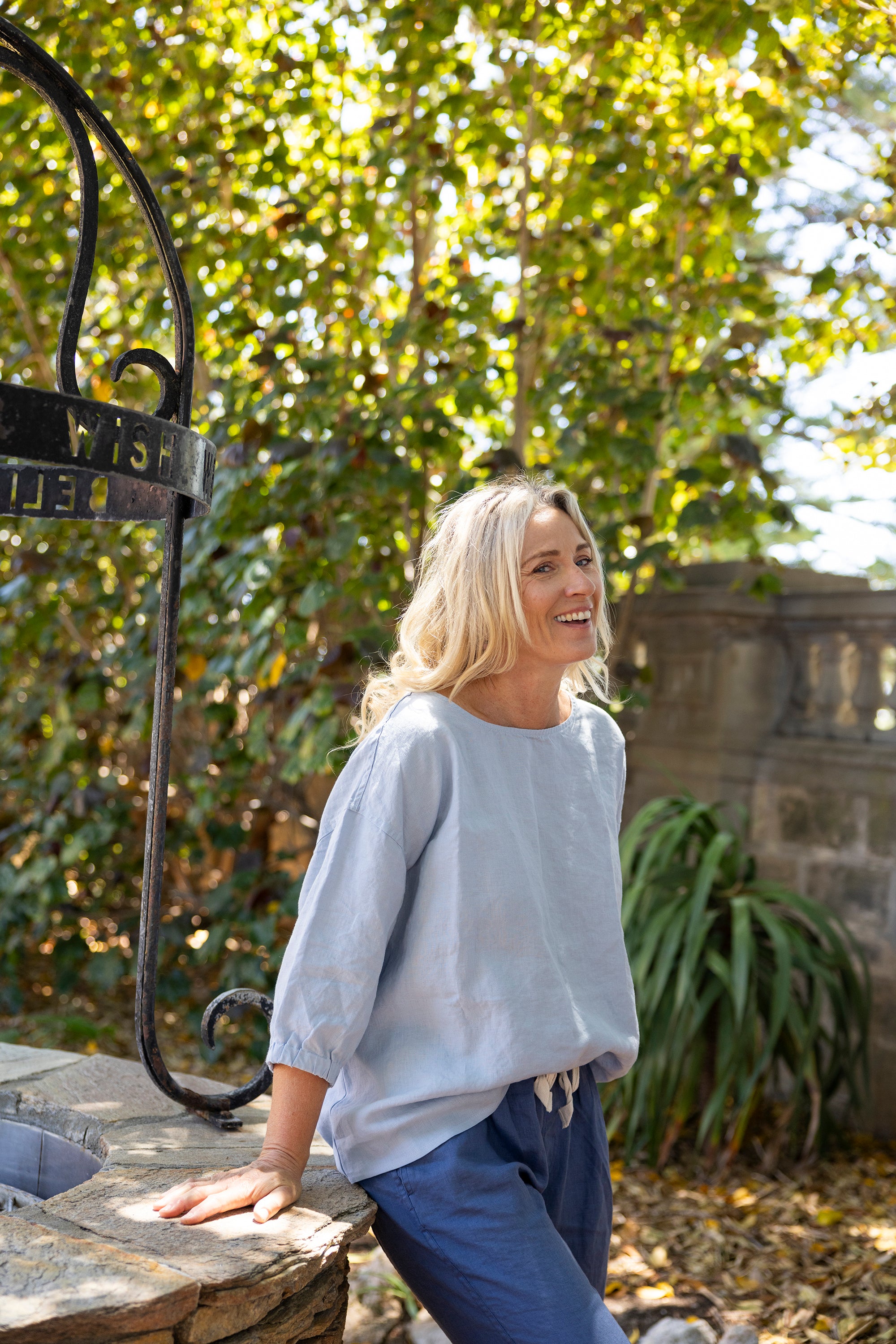 Woman standing outdoors wearing an oversized pale blue relaxed-fit boxy top paired with denim blue trousers with trees and a stone wall in the background