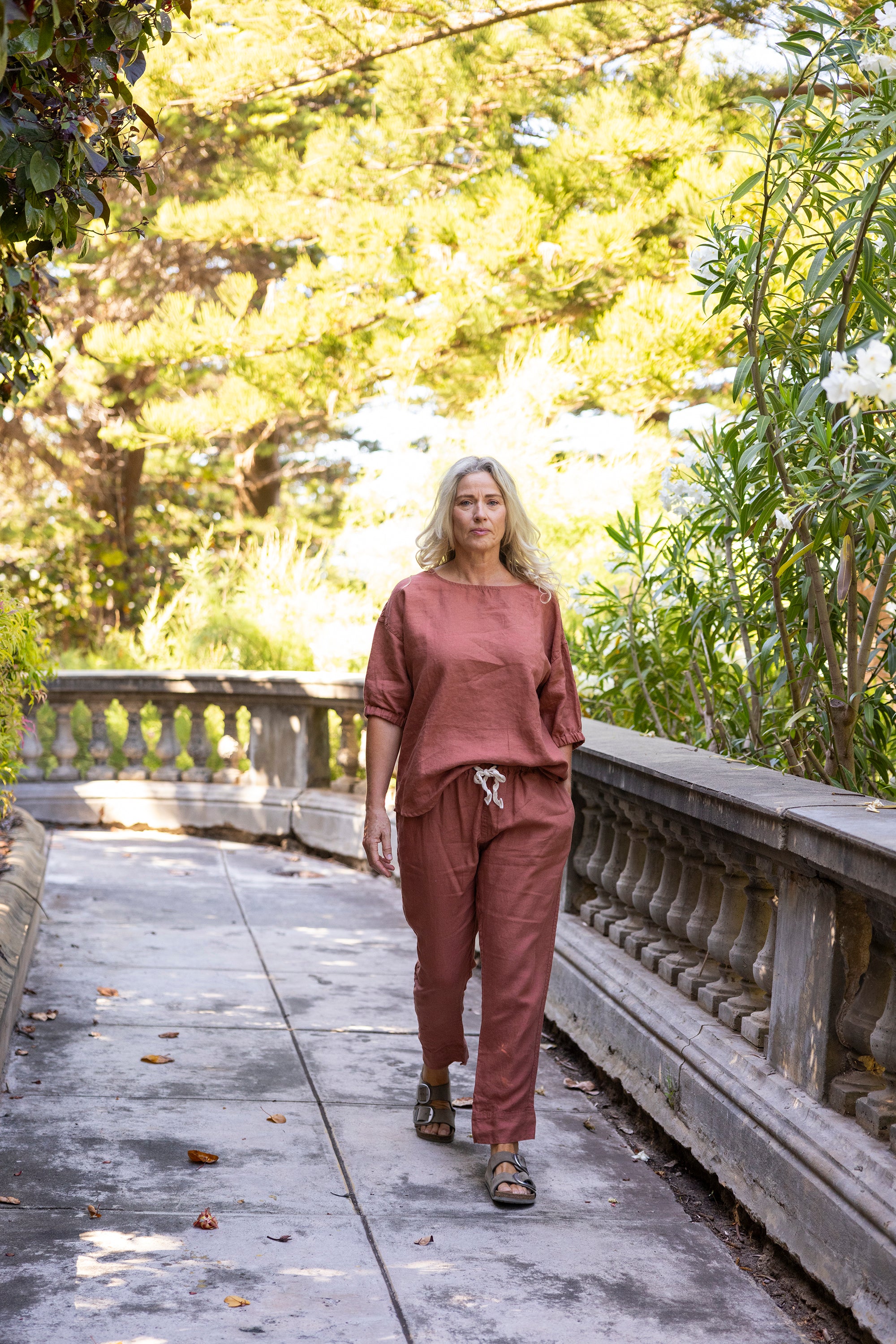 Woman in a rust outfit of oversized relaxed-fit top and straight leg trousers walking on a path with greenery around
