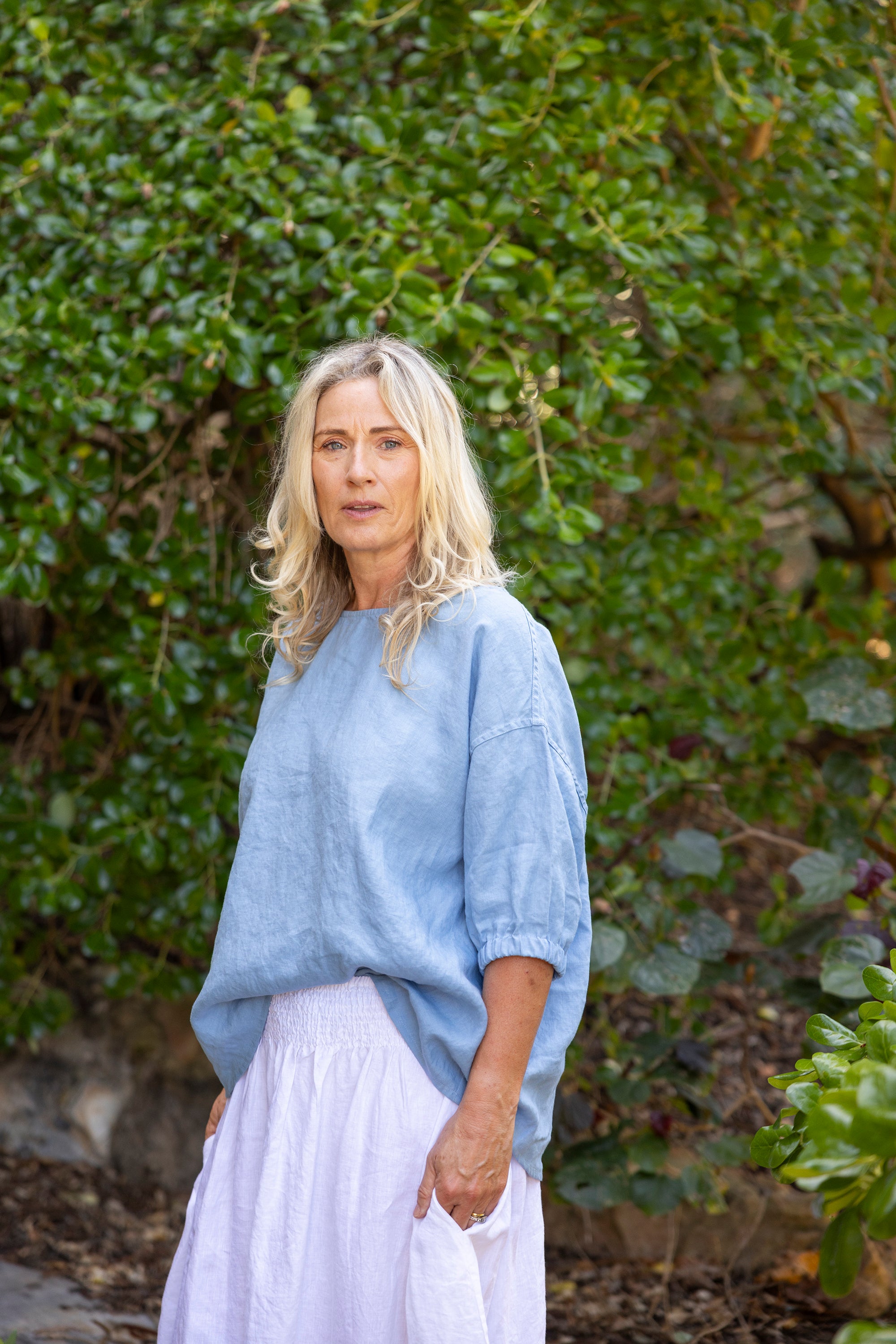 Woman wearing a light blue boxy top and white pants standing in front of green foliage.
