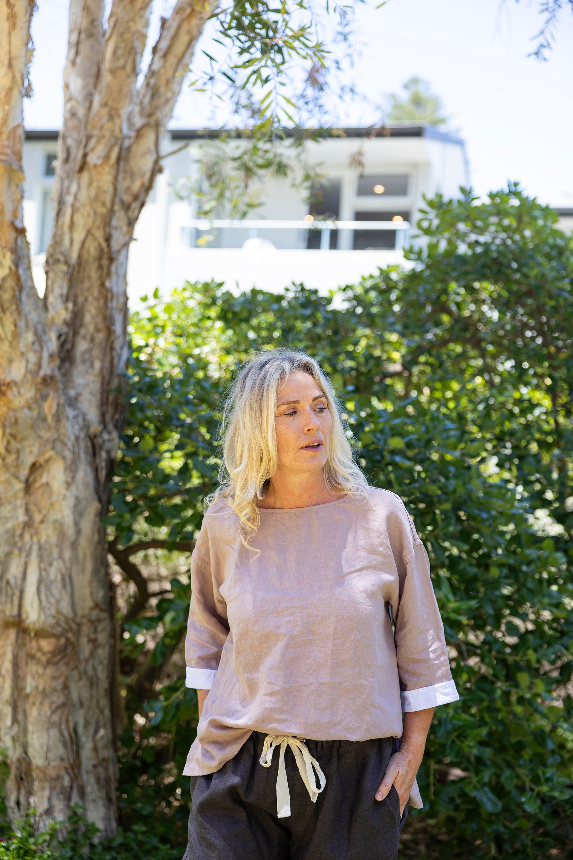 Woman wearing an oversized boxy top with white cuffs in a dusky rose colour outdoors in a garden with trees and a building in the background