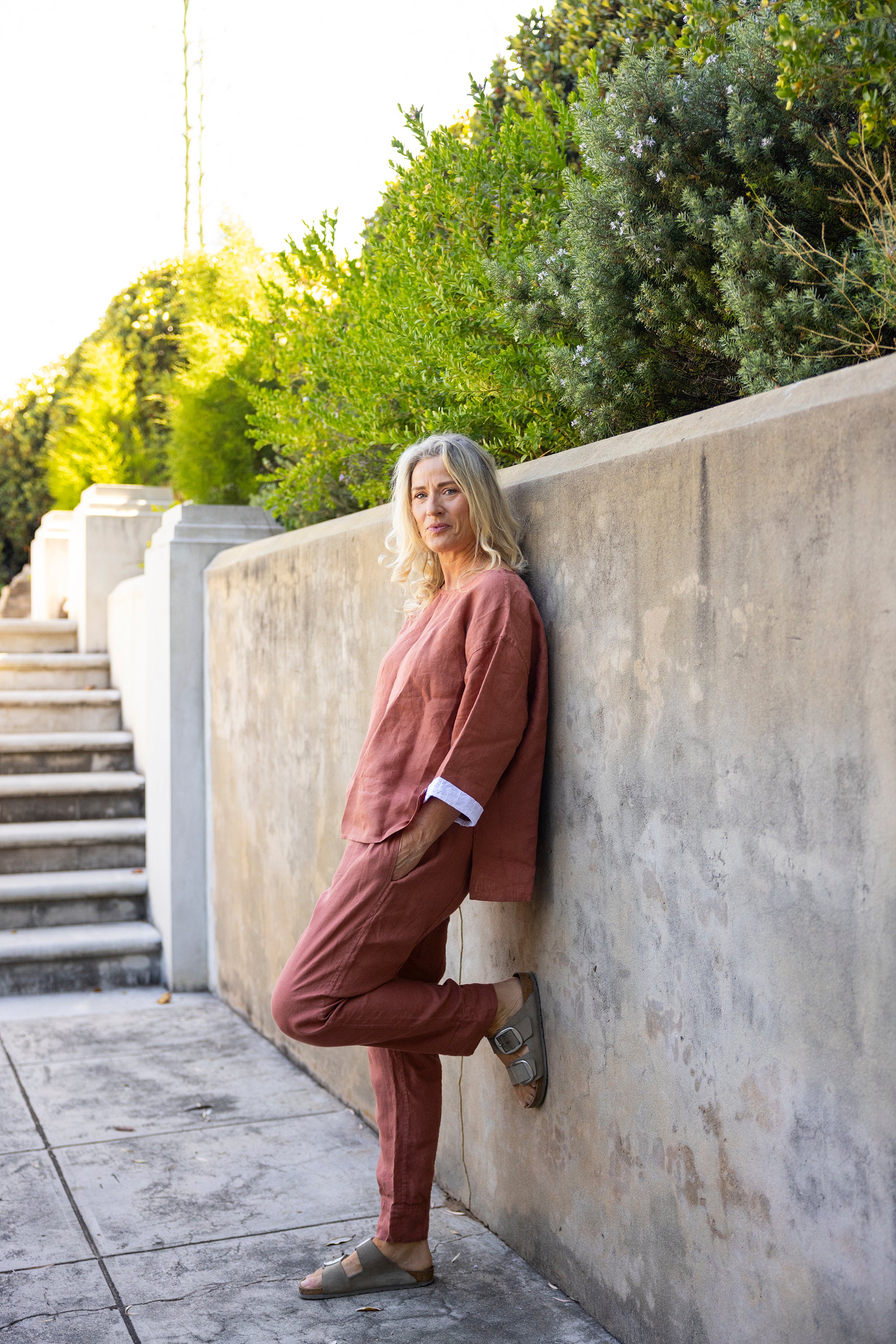 Woman in an oversized boxy top in a rust colour and matching pants against a stone wall with greenery in the background 
