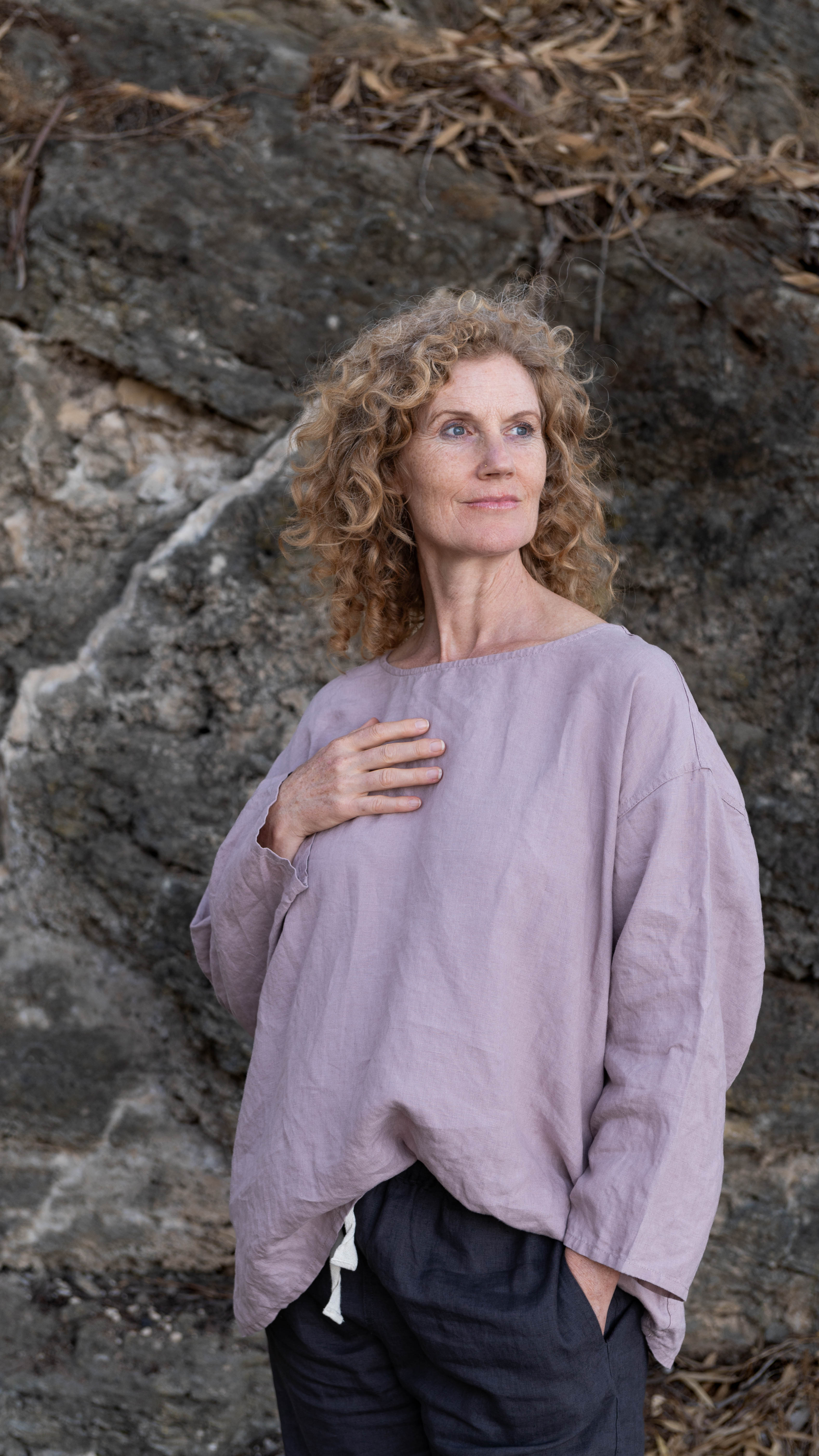 A woman standing outdoors wearing a relaxed fit, long-sleeved Boxy Top in a dusky pink color, paired with dark pants.