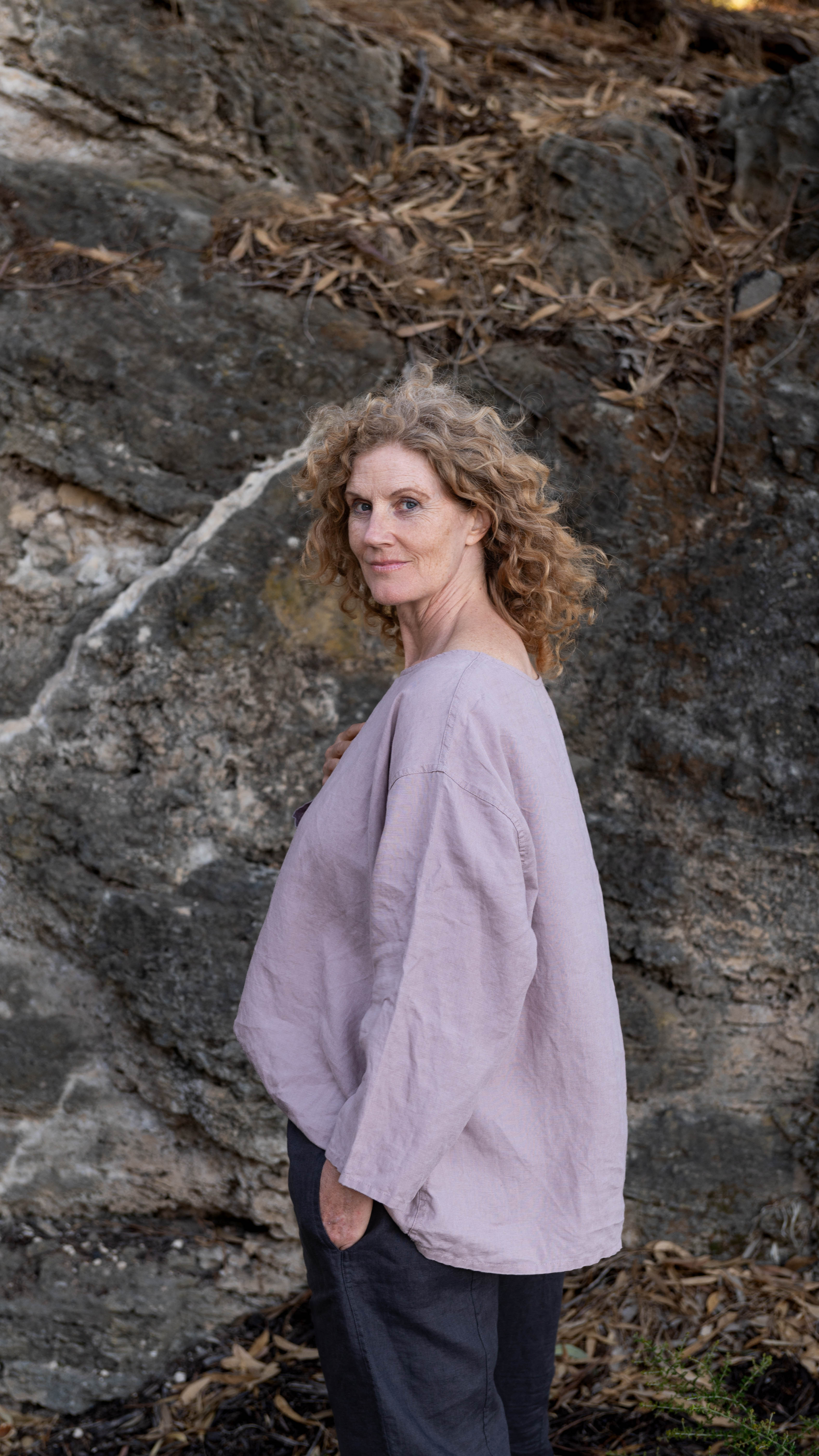 A woman standing outdoors wearing a relaxed fit, long-sleeved Boxy Top in a dusky pink color, paired with dark pants.