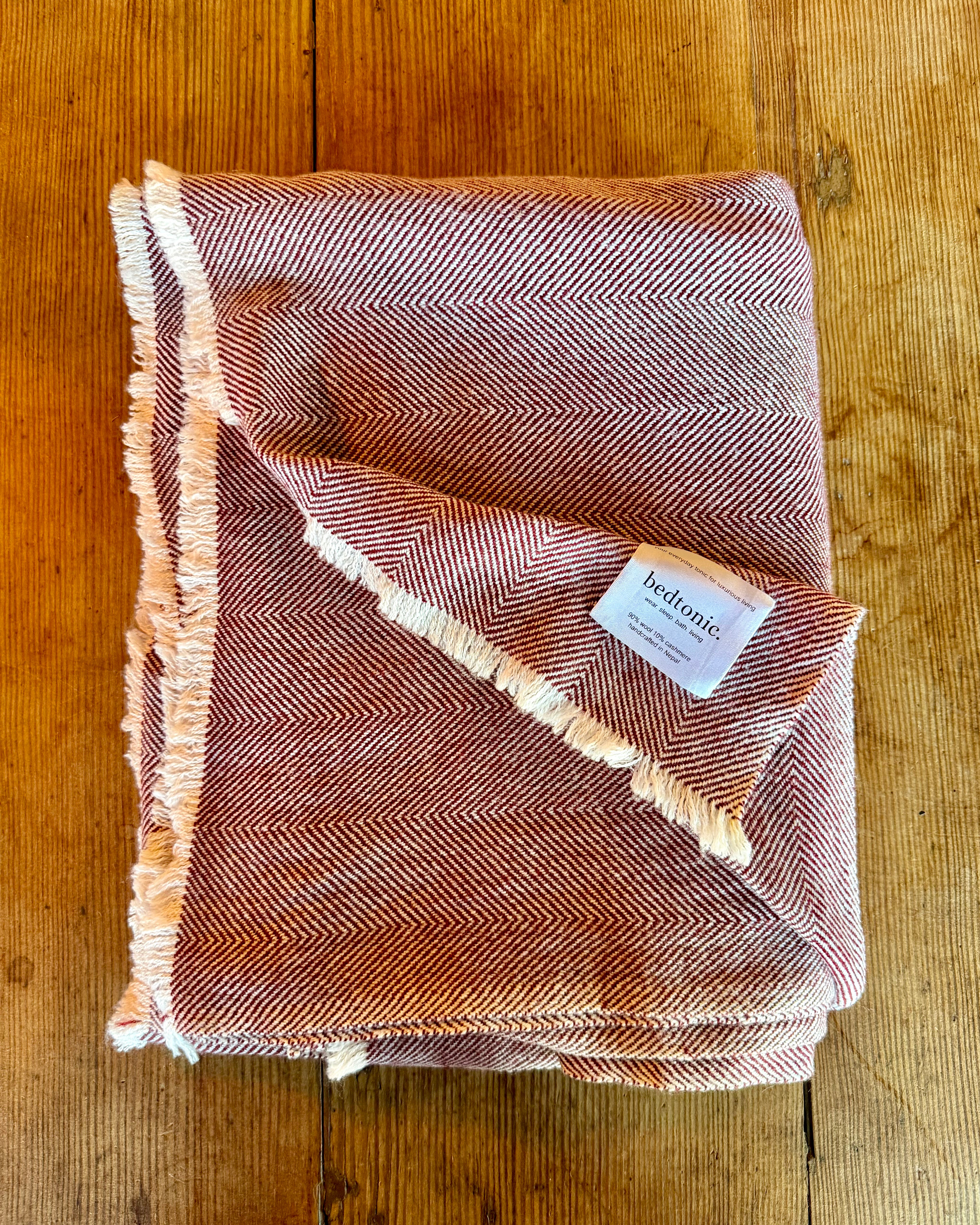 A folded woven throw blanket with a herringbone pattern in dusty rose and cream, featuring fringed edges and a white 'bedonic' brand tag, laid flat on a rustic wooden surface.