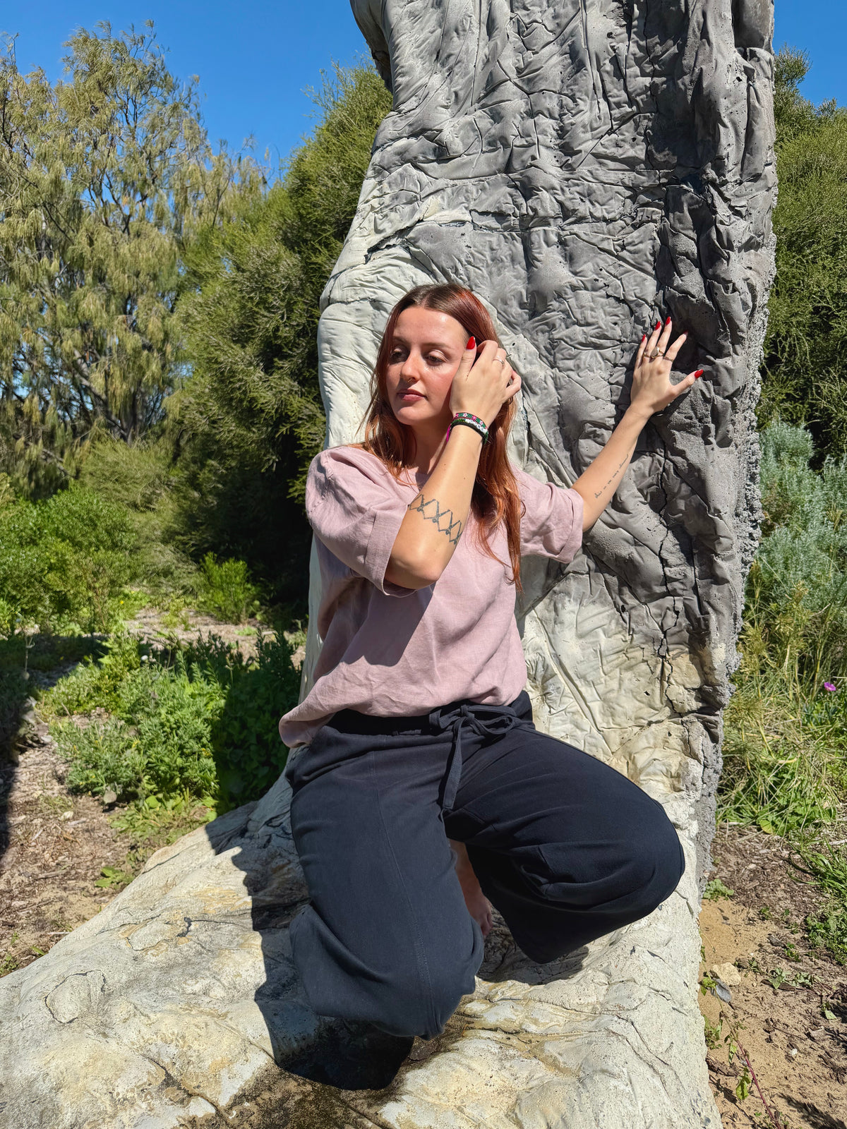 A woman kneels on a large rock in an outdoor garden setting, wearing a dusty pink short-sleeve top and dark navy wide-leg pants against a textured gray rock face.