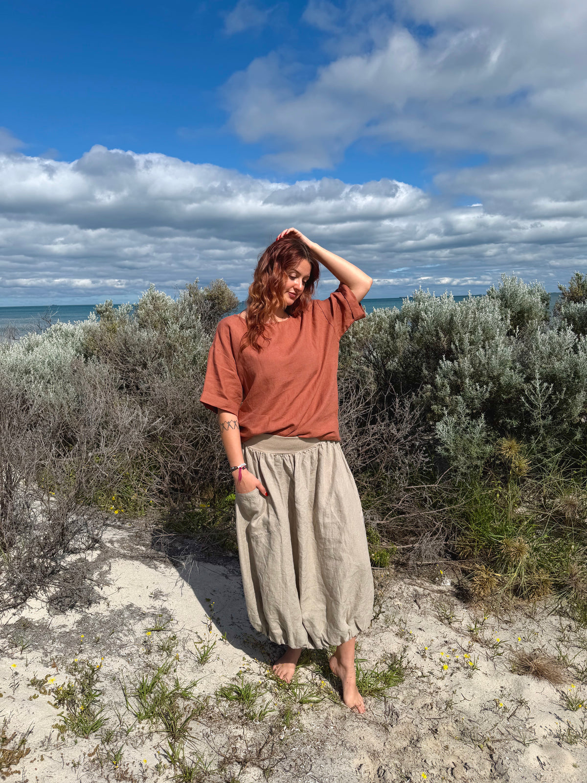 A woman stands outdoors on sandy ground with shrubs and the ocean under a blue sky, wearing a rust-orange short-sleeve top and a loose beige skirt.