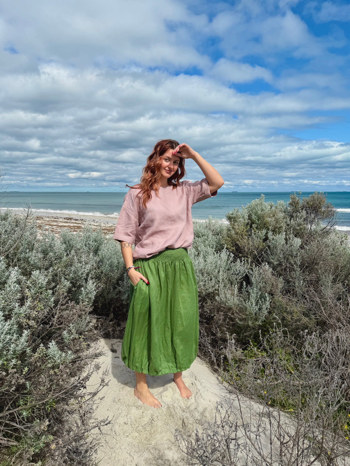 A woman stands barefoot on a sandy path between green shrubs, wearing a dusty pink short-sleeve top and a flowing olive green skirt, with the ocean and cloudy blue sky behind her.