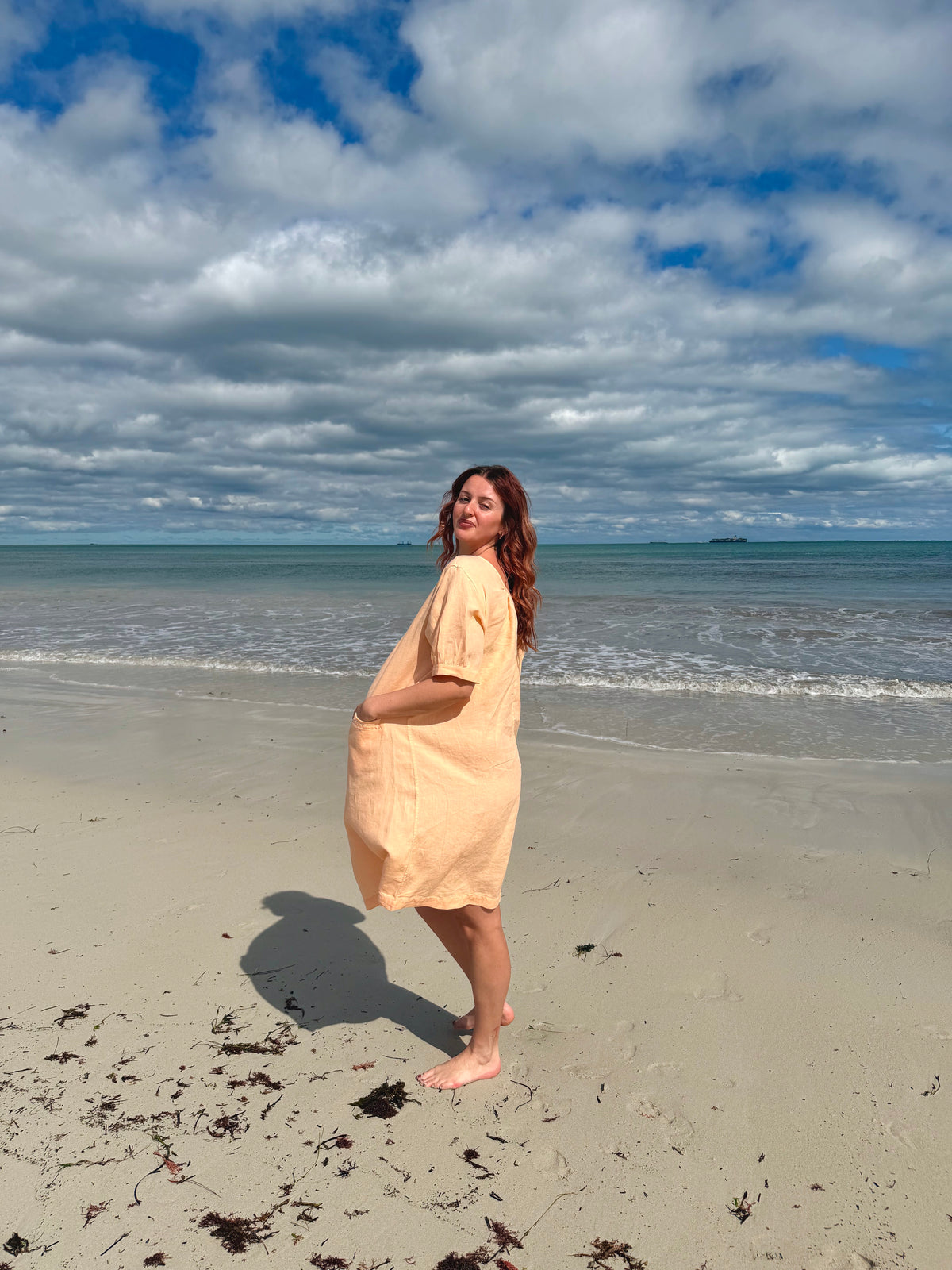 A woman stands barefoot on a sandy beach wearing a light peach-colored short-sleeve tunic dress, looking back over her shoulder under a cloudy blue sky.