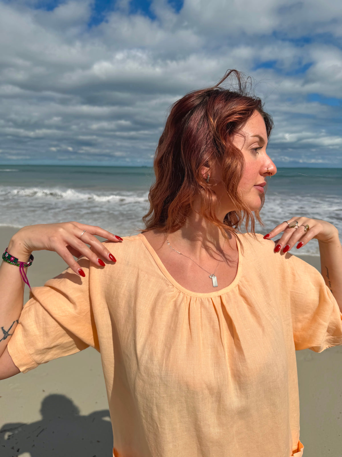 A woman with reddish-brown hair stands on a beach wearing a light peach-colored short-sleeve top, looking to the side with the ocean and cloudy sky in the background.