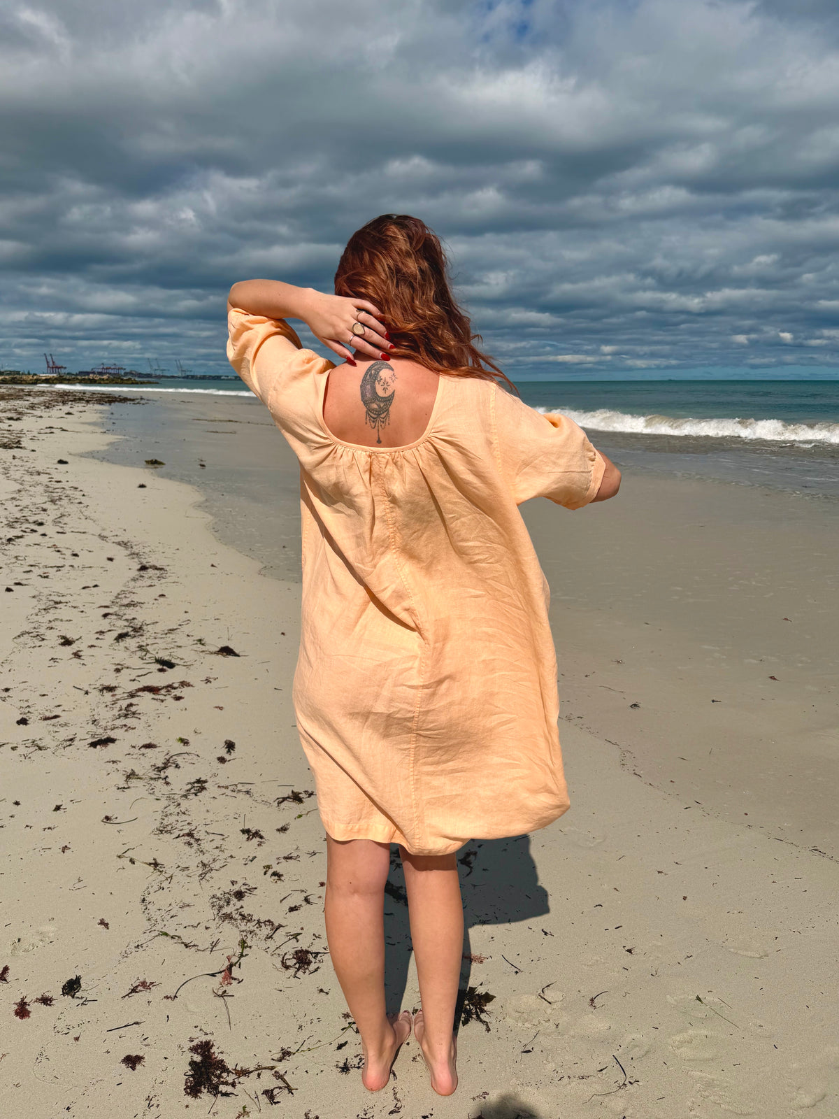 A woman stands on a sandy beach facing the ocean, wearing a loose, short-sleeve peach-colored linen dress. The dress has a gathered back neckline with a small butterfly motif and is shown in a lifestyle outdoor setting under a cloudy sky.