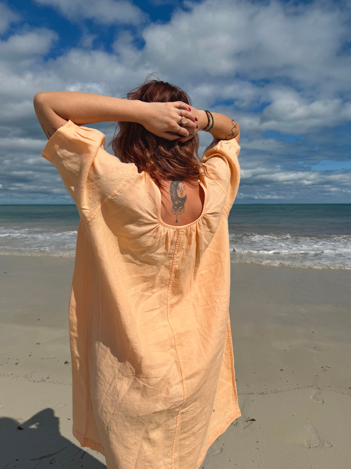 A woman stands on a sandy beach with her back to the camera, wearing a loose, peach-colored linen cover-up dress. The ocean and a cloudy blue sky are visible in the background.