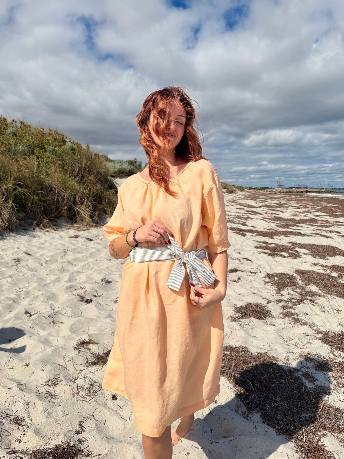 A woman with red hair stands on a sandy beach wearing a peach-colored linen dress with a light gray fabric tie at the waist, with dunes and a cloudy sky in the background.