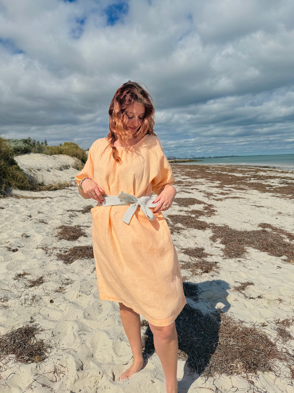 A woman wearing a peach-colored bathrobe stands on a sandy beach with the ocean and cloudy sky in the background.
