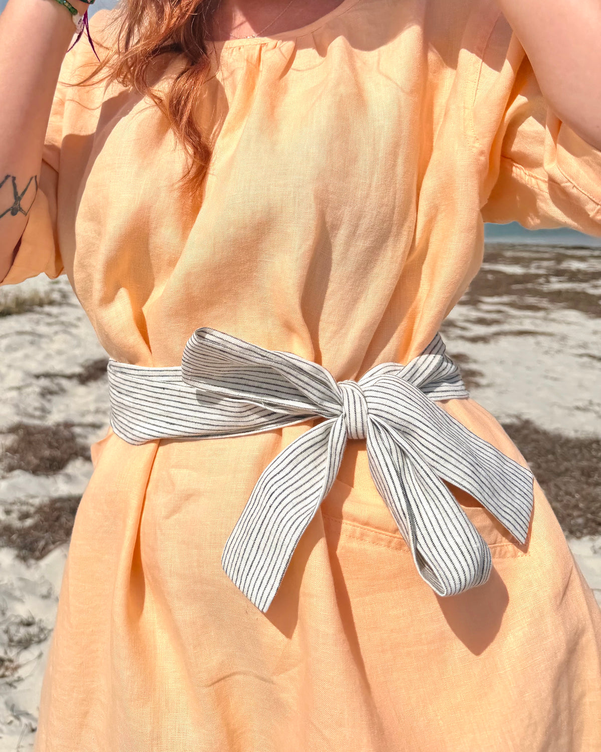 A close-up of a person outdoors wearing a peach-colored linen dress with a wide gray and white striped fabric tie belt knotted at the waist.