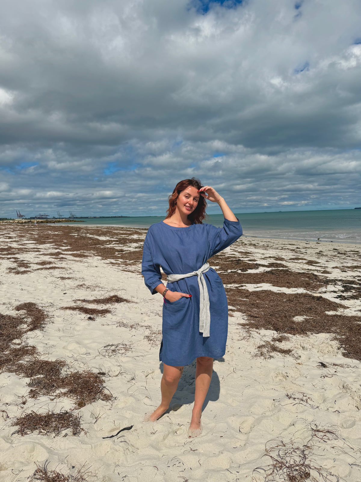 A woman stands on a sandy beach wearing a blue long-sleeve dress with a white tie belt. The background shows a cloudy sky, ocean, and shoreline with patches of seaweed.