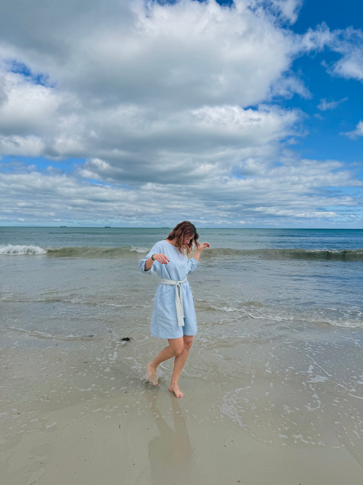 A woman in a light blue bathrobe walks barefoot on a sandy beach with gentle waves under a blue sky with large white clouds.