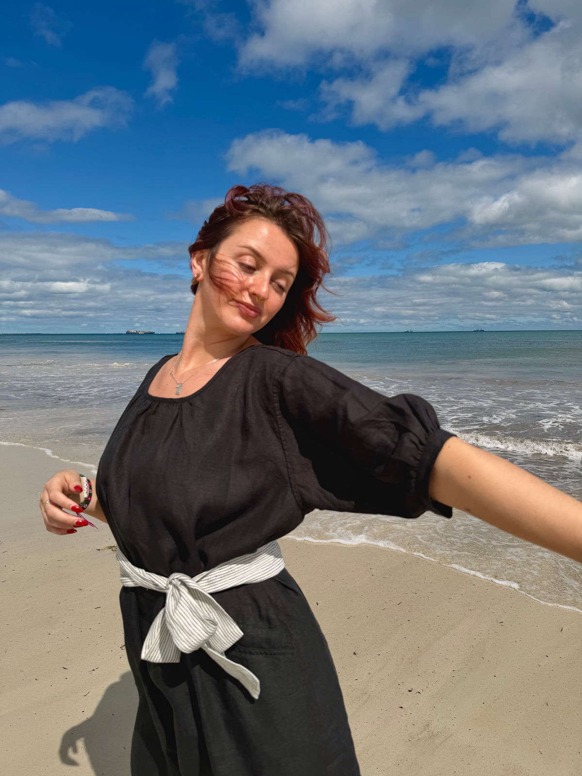 A woman posing on a sandy beach wearing a black dress with a white striped waist tie, with the blue ocean and cloudy sky in the background.