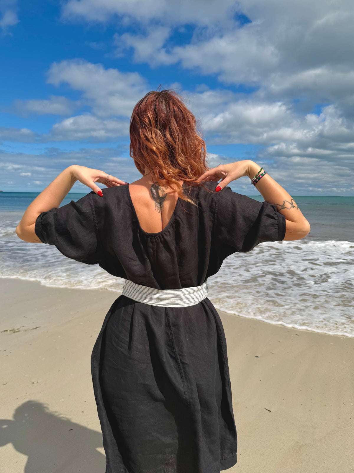 A woman with reddish-brown hair stands on a sandy beach with the ocean and blue sky behind her, wearing a loose black dress with a white sash at the waist. She is seen from behind, adjusting her hair with both hands.
