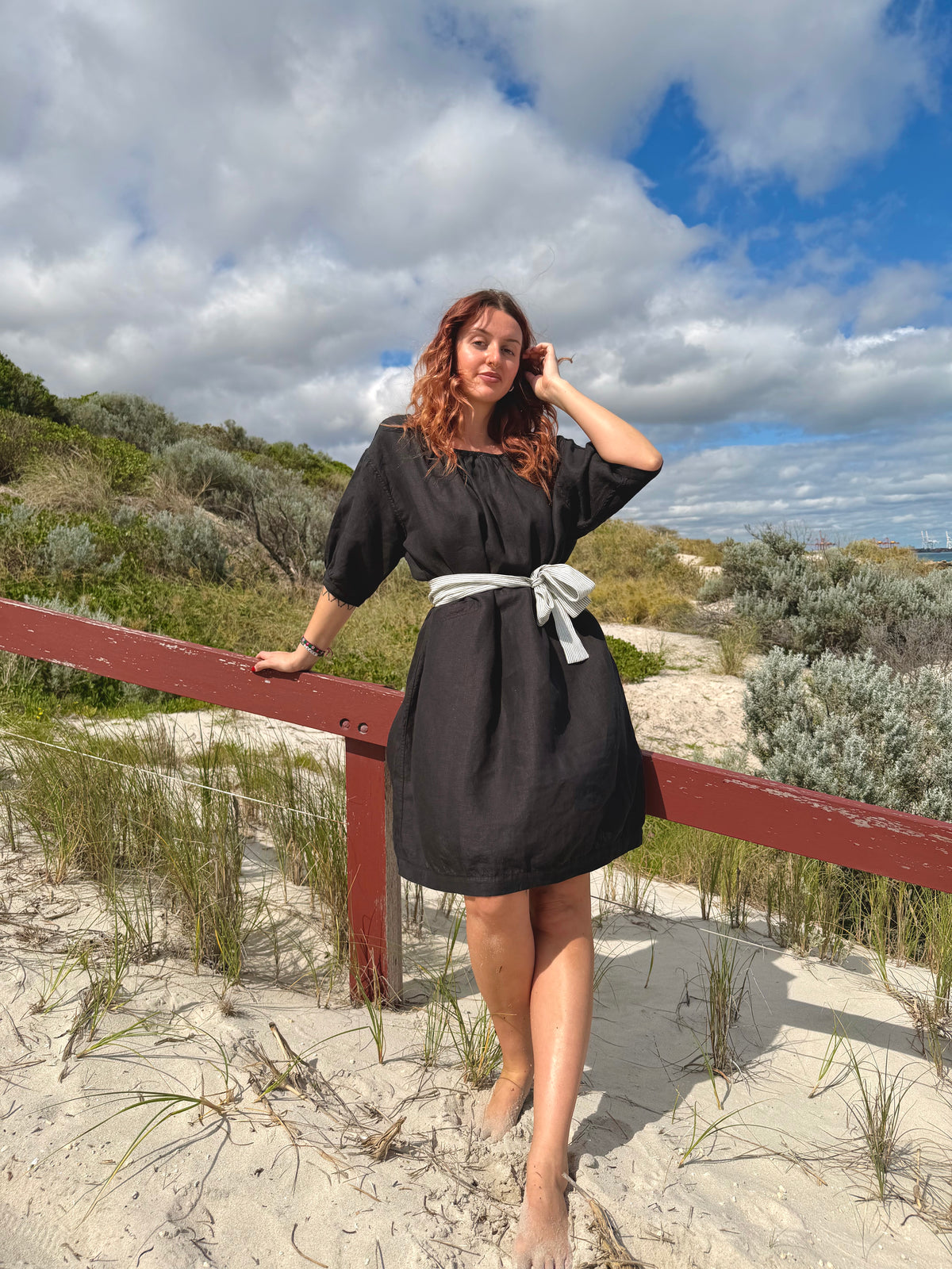 A woman wearing a black dress with a white waist tie stands on a sandy beach dune next to a wooden railing, with dune grass, shrubs, and a cloudy blue sky in the background.