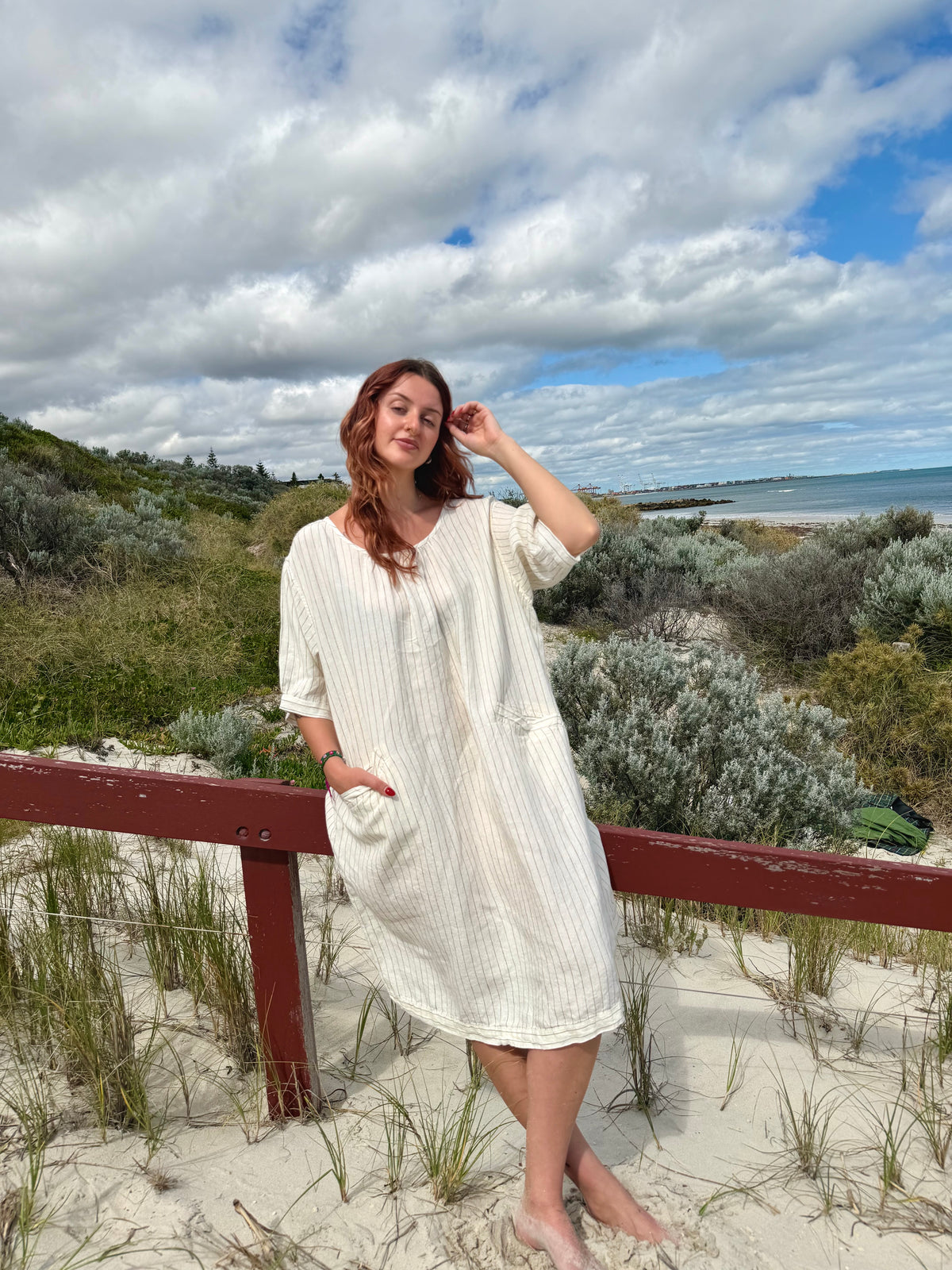 A woman wearing a cream-colored, short-sleeved ribbed linen dress stands on a sandy dune with a wooden railing, looking towards the camera with a beach and cloudy sky in the background.