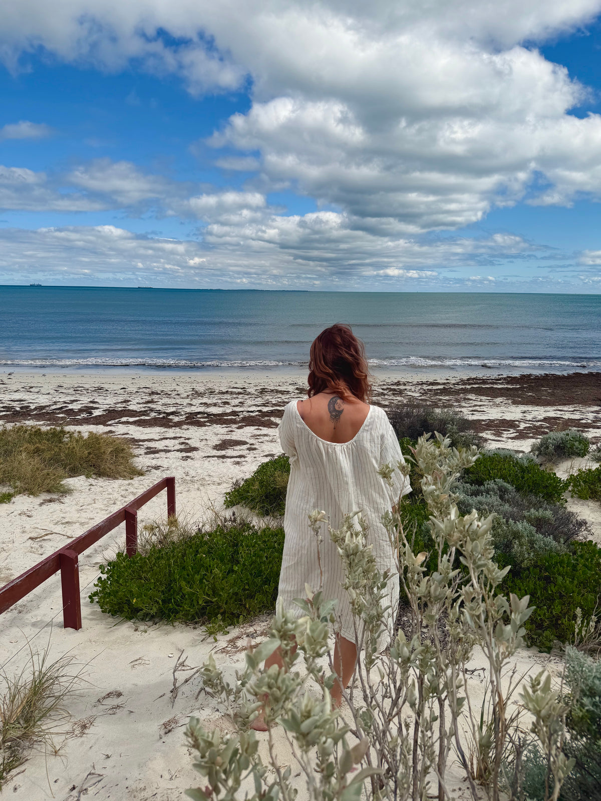 A woman stands on a sandy beach dune facing the ocean, wearing a loose white backless cover-up dress. The scene includes green beach vegetation, a wooden railing, and a blue sky filled with large white clouds.