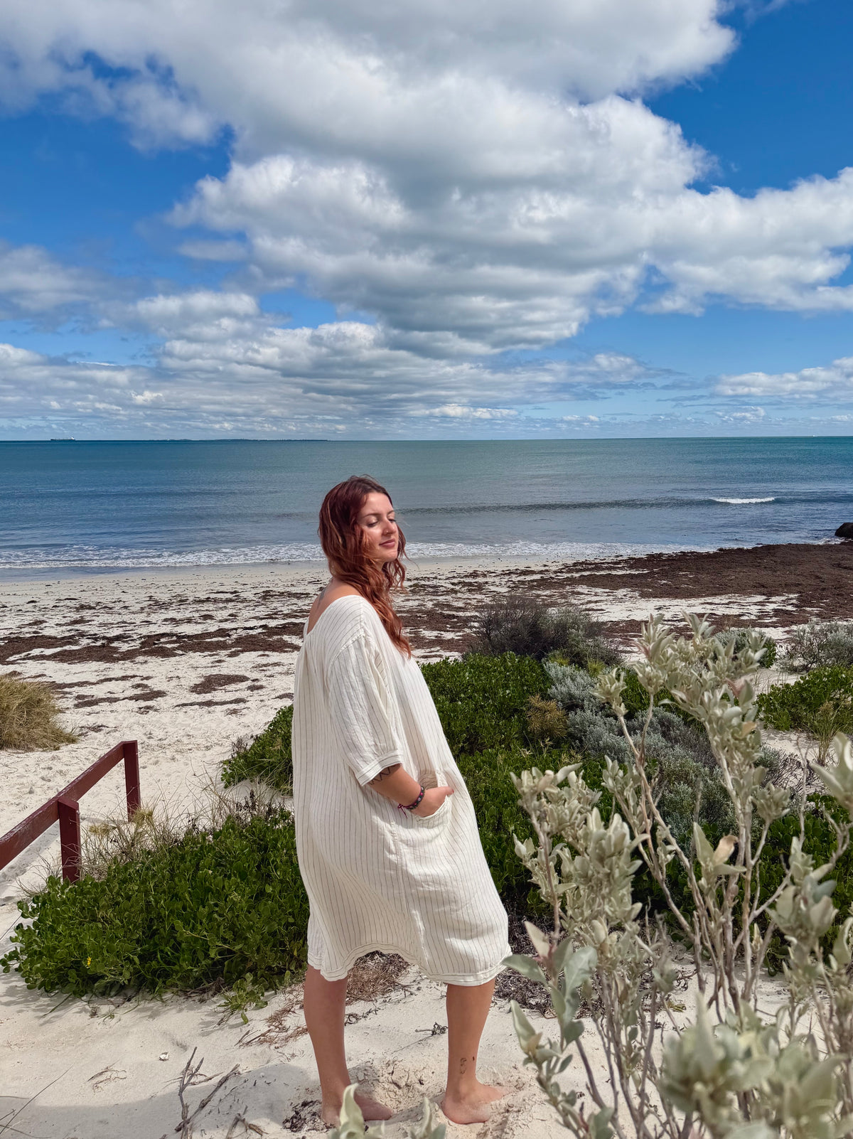A woman wearing a cream ribbed knit short-sleeve dress stands on a sandy beach with ocean, sky, and greenery in the background.