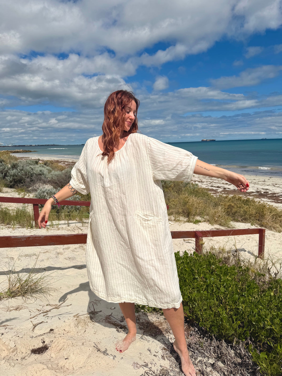 A woman wearing a cream-colored, vertically striped linen tunic dress stands on a sandy beach with the ocean and cloudy sky in the background.
